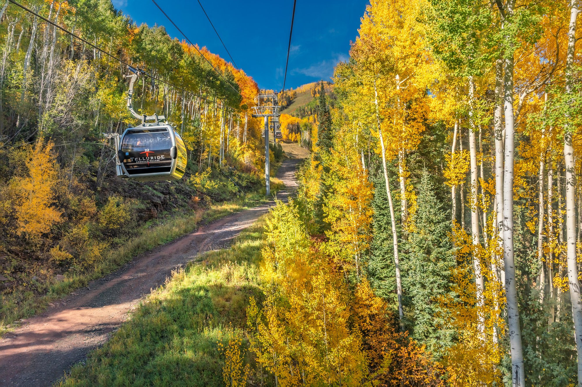 A scenic mountain landscape with a colorful forest and a dirt trail, featuring a gondola lift with several cabins, including one labeled 'Telluride'.