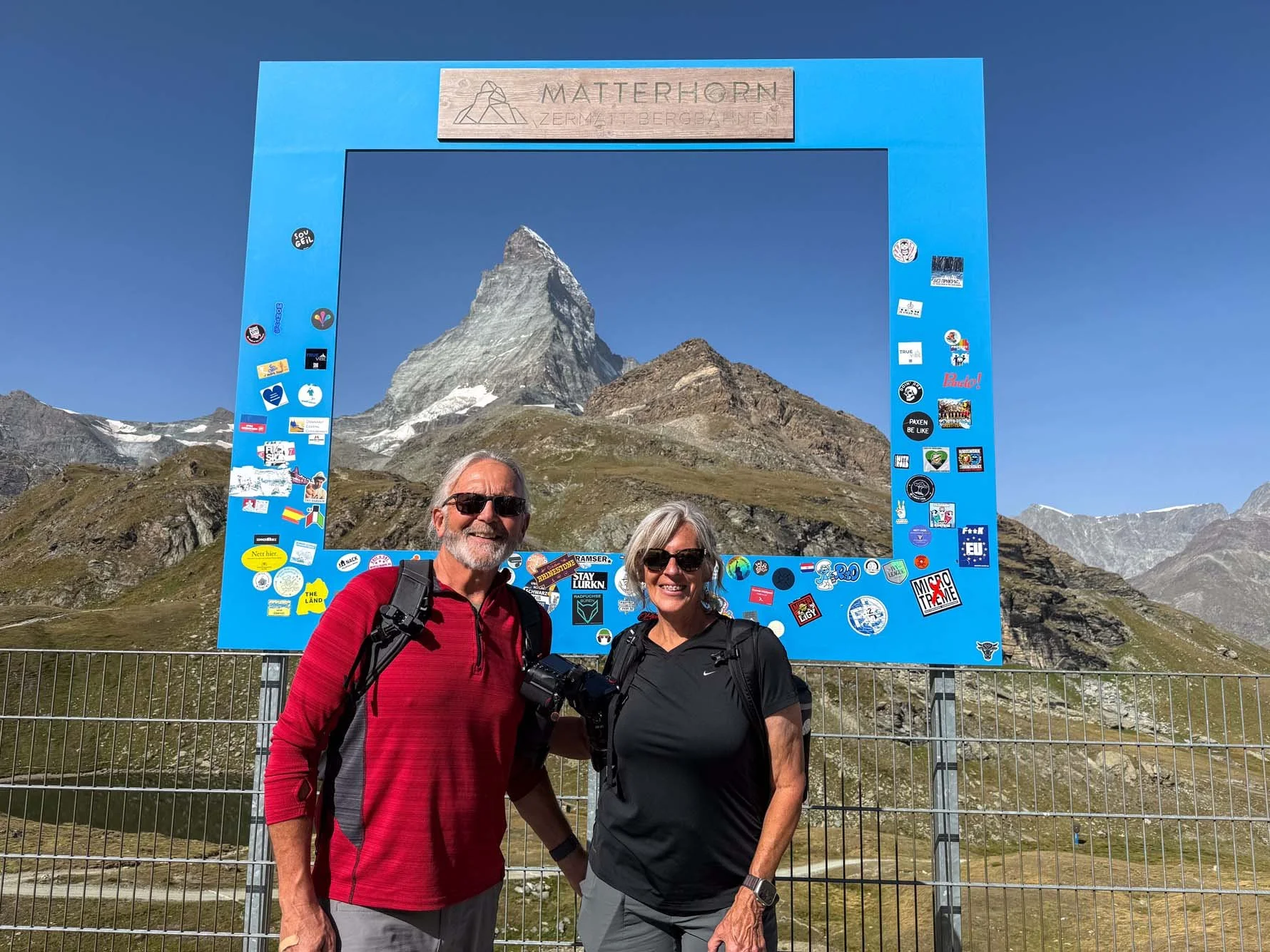 A smiling man and woman with backpacks standing in front of a large blue photo frame with the Matterhorn mountain in the background, surrounded by stickers.