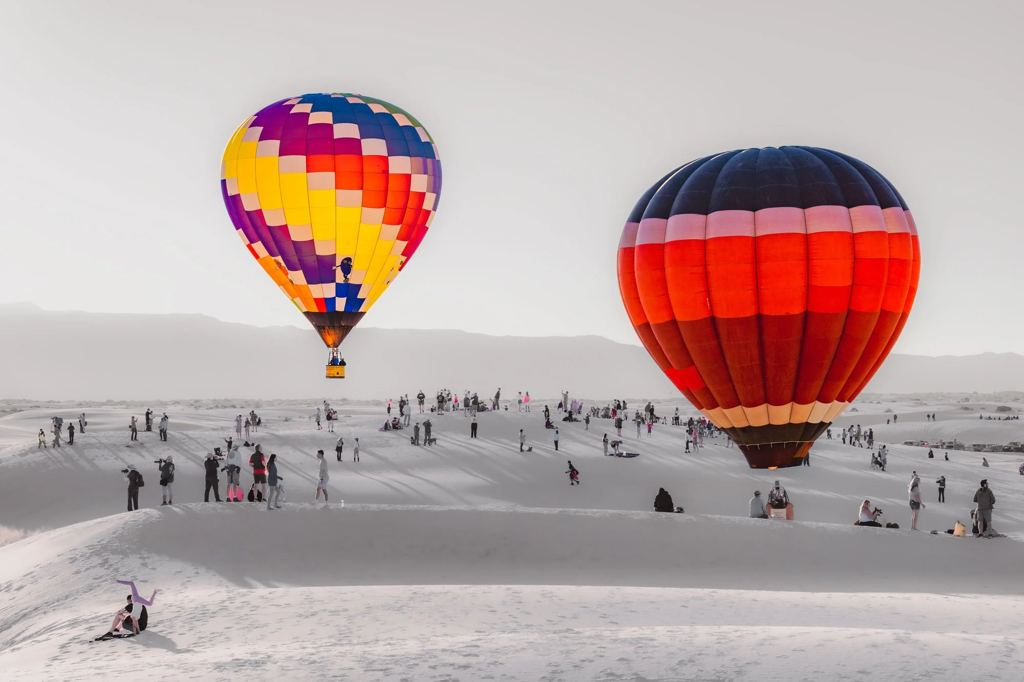 Two colorful hot air balloons floating above a sandy landscape with many people walking and taking photos.
