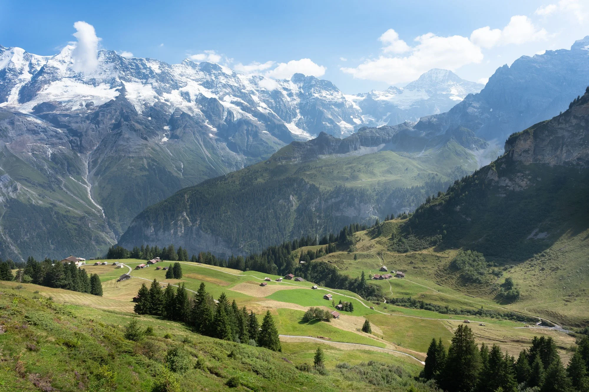A scenic view of lush green fields and small houses in a valley surrounded by tall, snow-capped mountains under a partly cloudy sky.