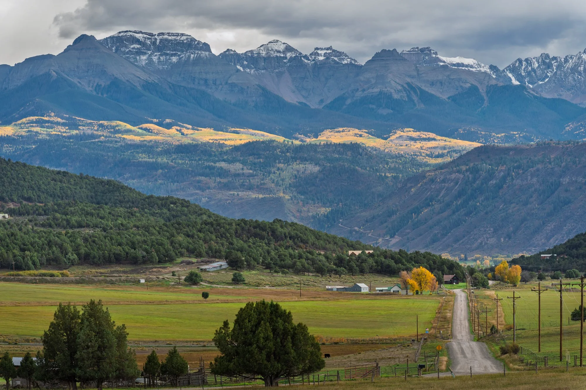 Picturesque landscape of lush green fields, winding dirt road, scattered farm buildings, dense forests, and towering snow-capped mountains under cloudy sky.