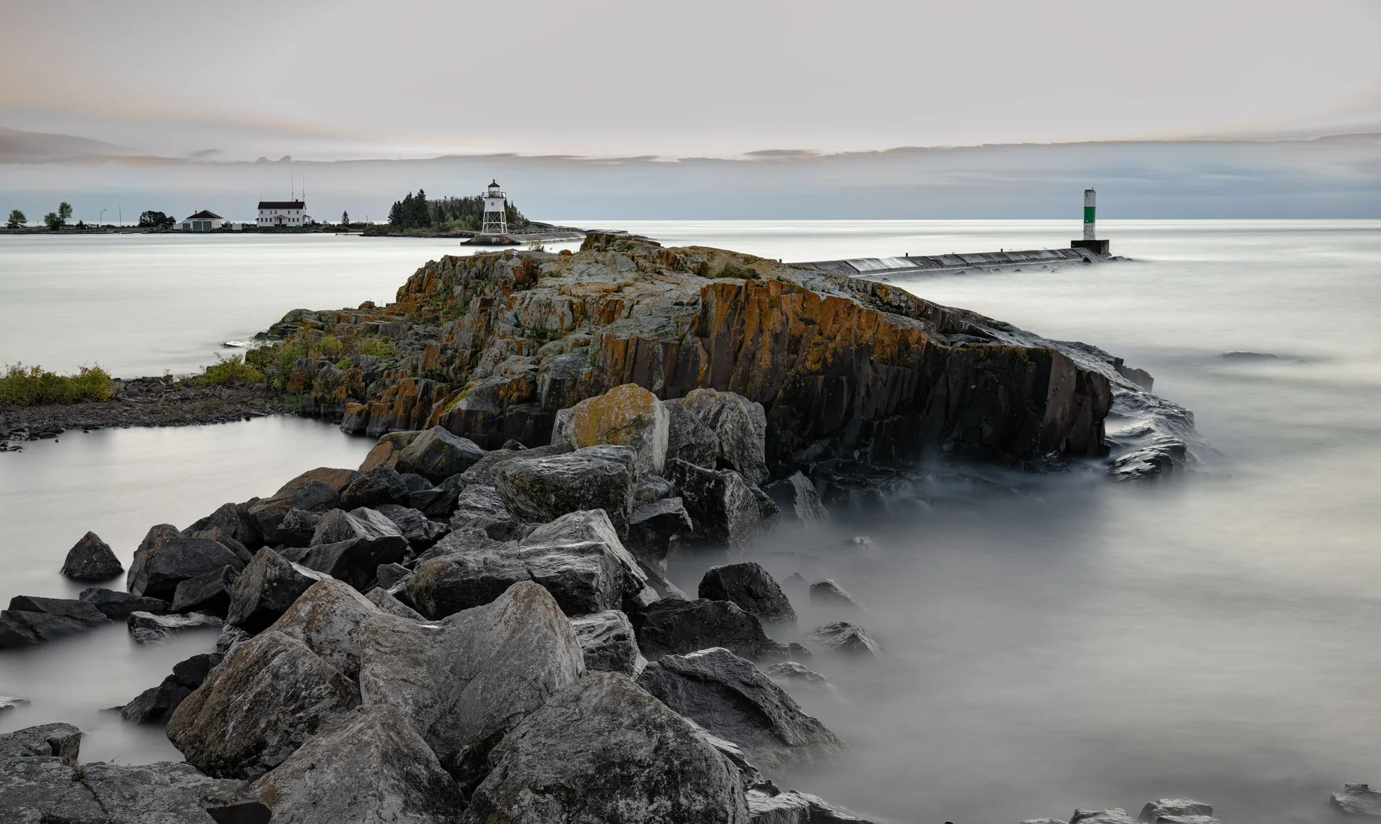 A rocky shoreline with a lighthouse, small building, and trees in the background, along with a breakwater extending into the water under an overcast sky.