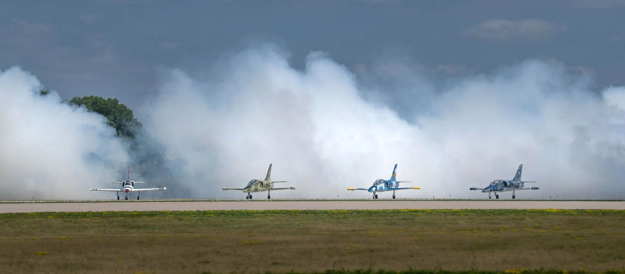 Four fighter jets parked on a runway with smoke or vapor clouds billowing behind them, and trees in the background.