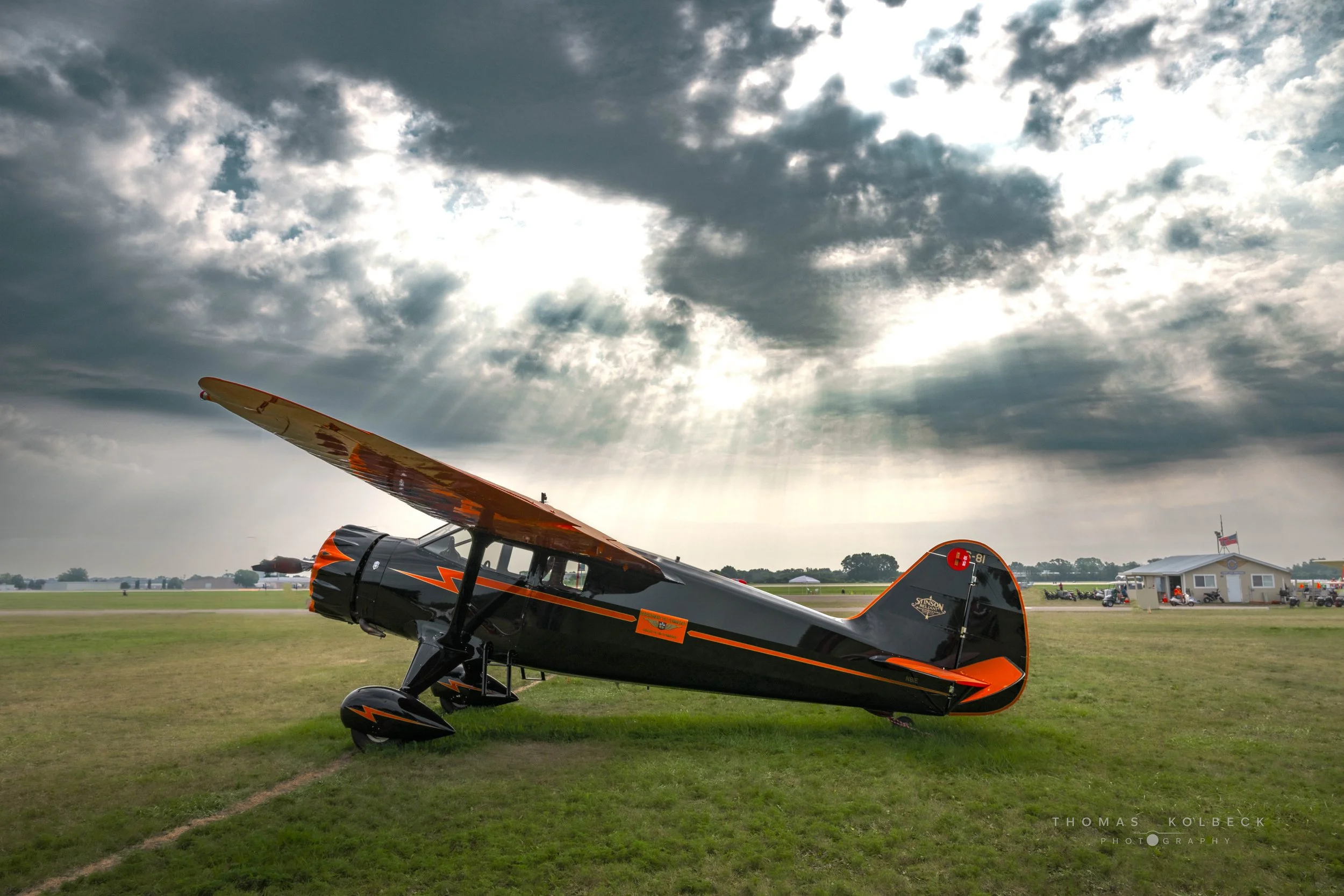 A vintage black and orange monoplane aircraft parked on a grass field under a cloudy sky with rays of sunlight breaking through.