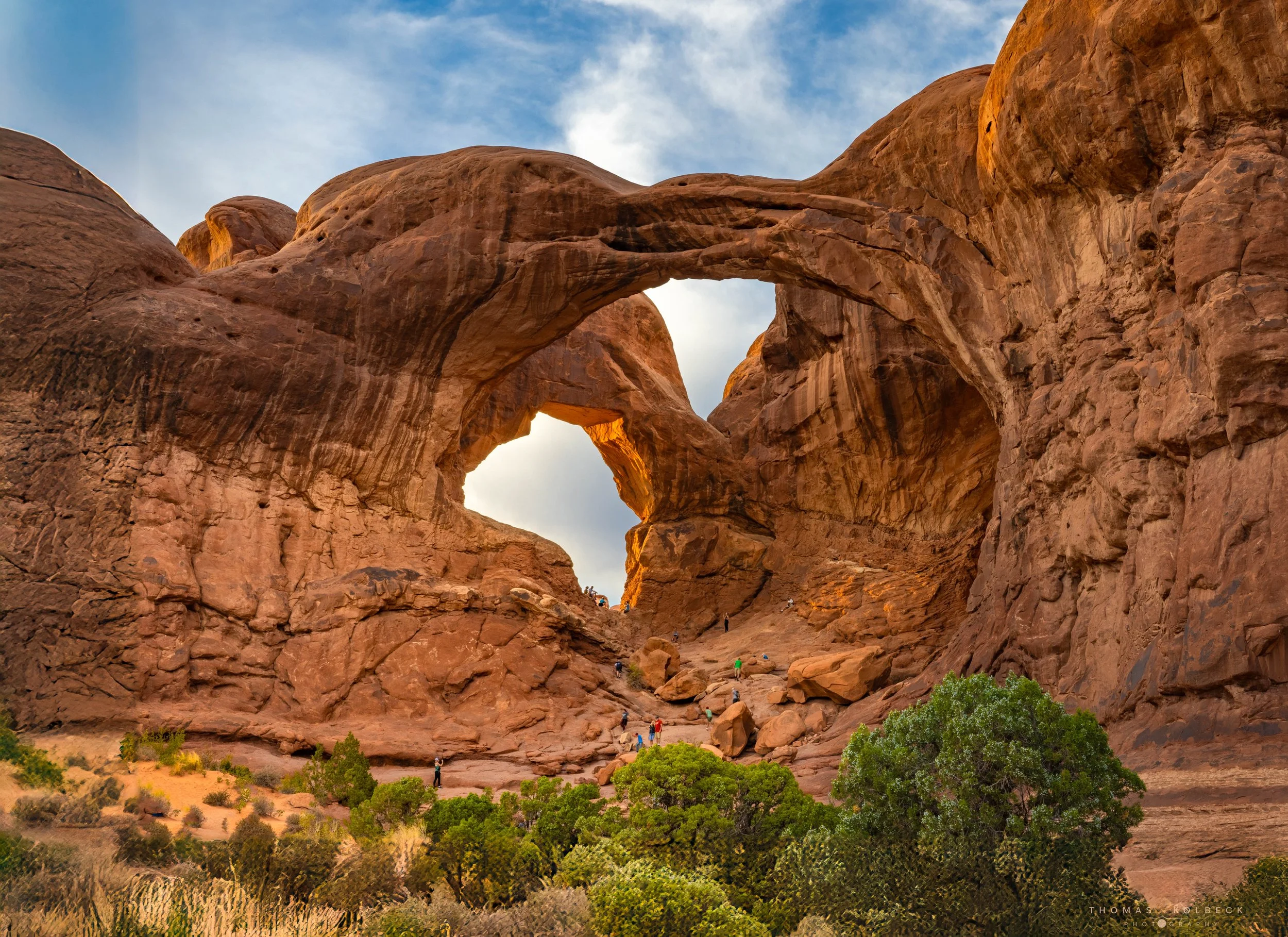 Red rock formations featuring a natural double arch in a desert landscape with green bushes in the foreground and a blue sky with clouds above.