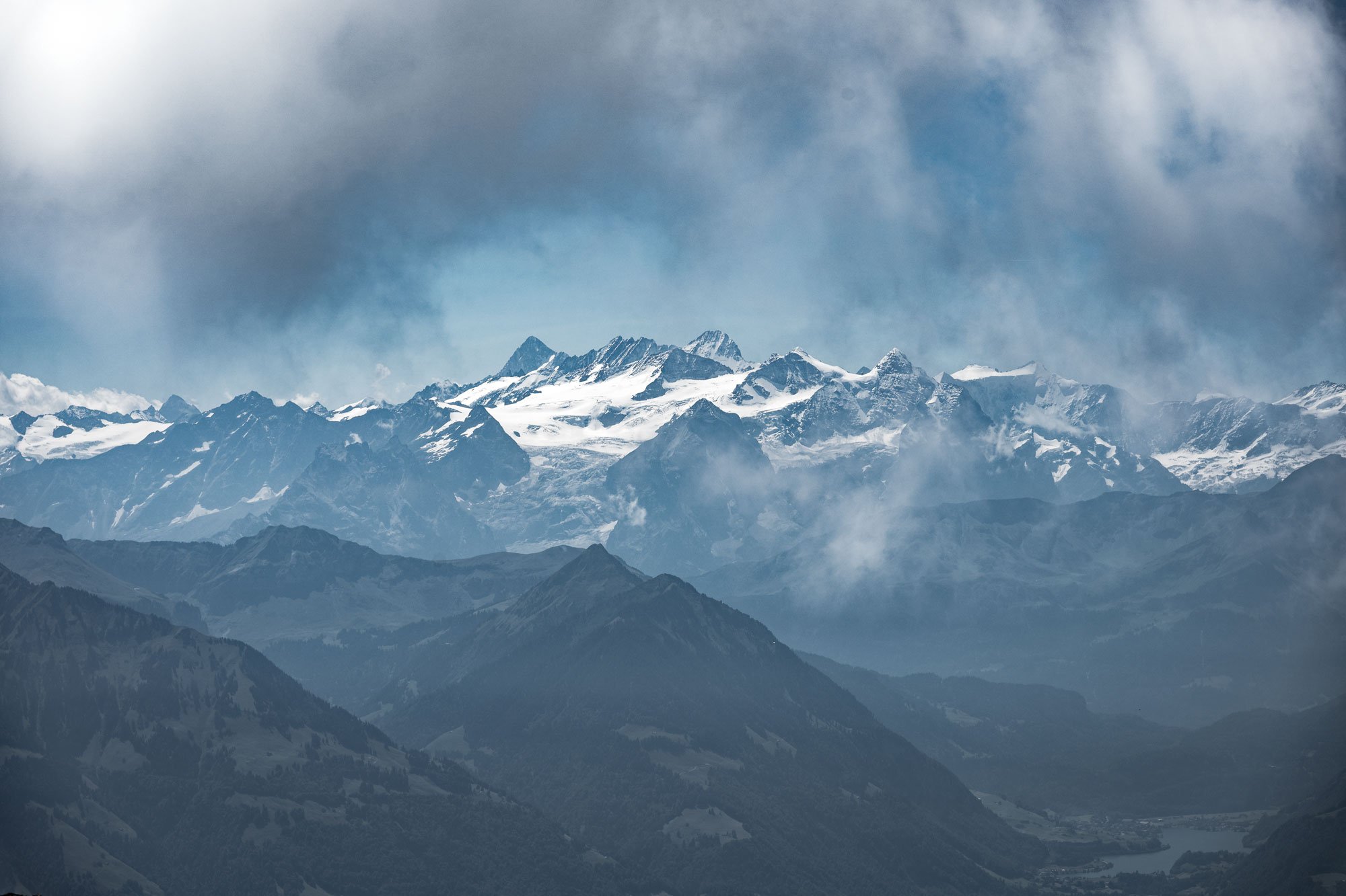 Snow-capped mountains with a cloudy sky in the background and green hills and a river in the foreground.