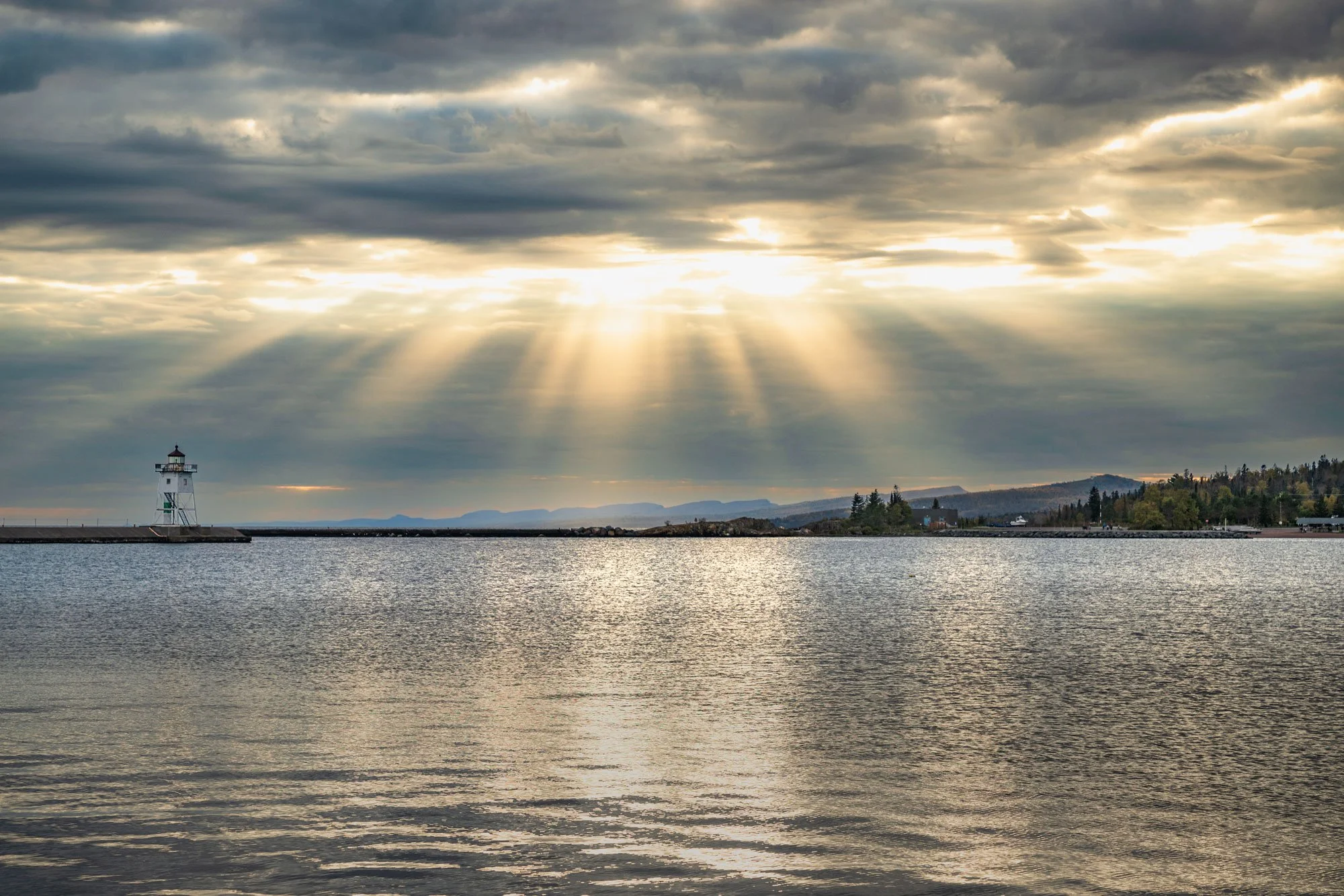 Sun rays breaking through cloud cover over a body of water with a lighthouse on the pier and distant mountains.