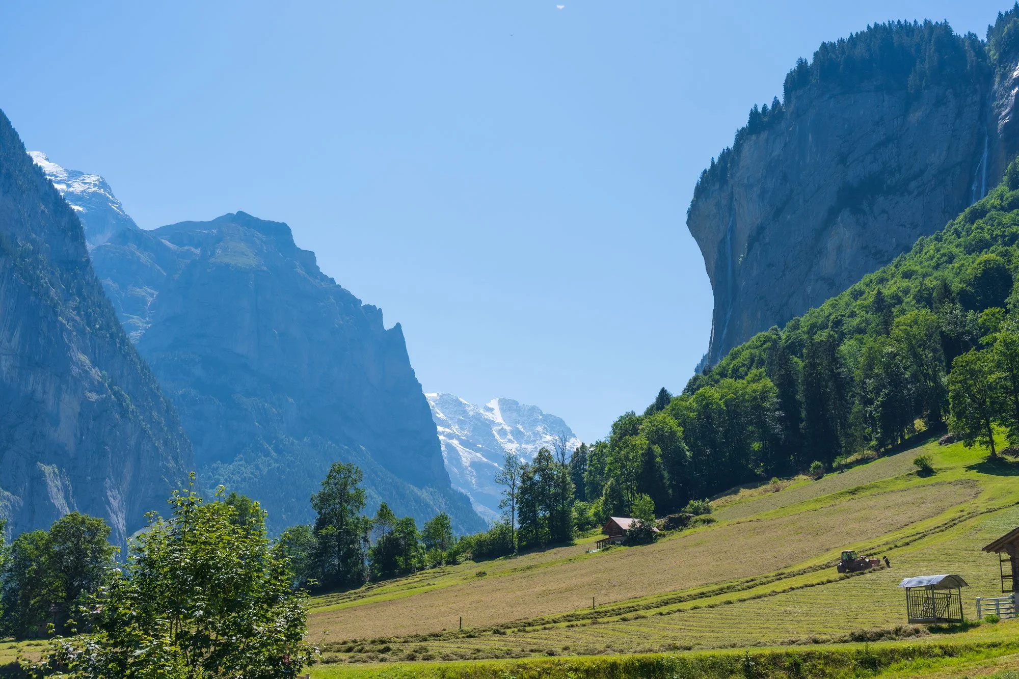 A scenic landscape featuring mountain cliffs, snow-capped peaks, green forested hills, and a cultivated field with farm buildings and a tractor under a clear blue sky.