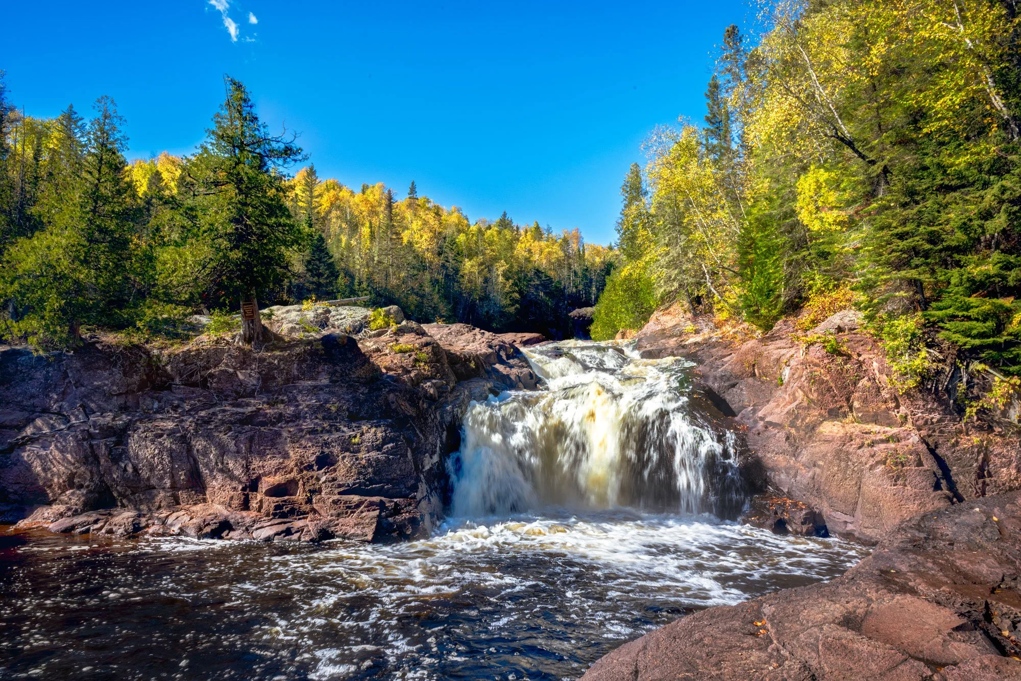 A waterfall flowing over rocks in a forest with green and yellow leaves under a clear blue sky.