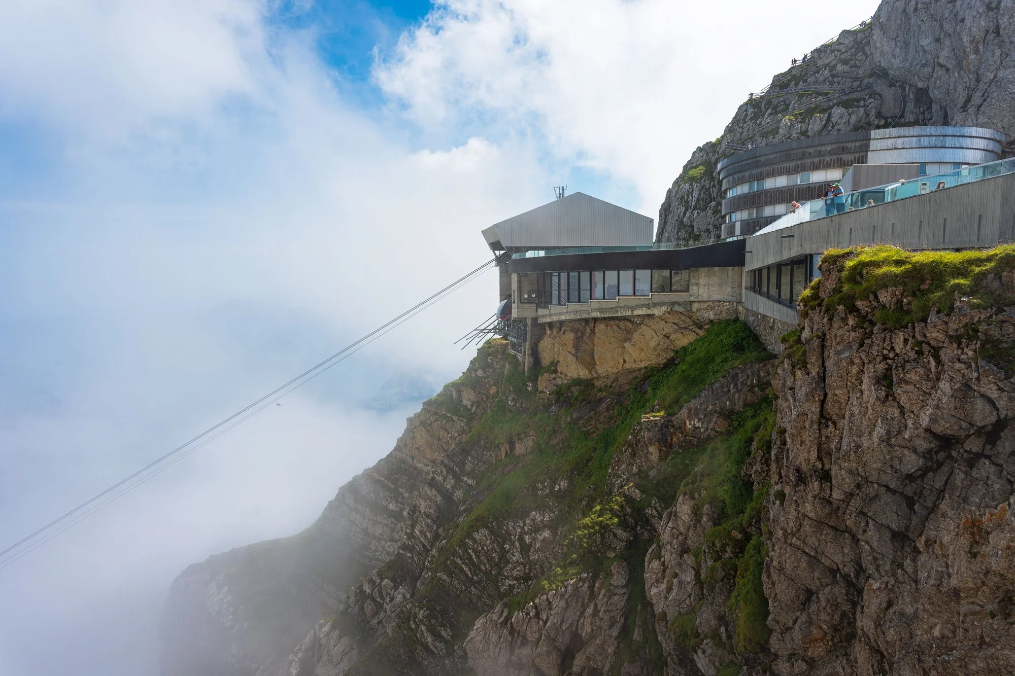 A mountain hotel building on a steep cliff, with glass balconies and cable cars passing nearby, surrounded by clouds and rocky terrain.