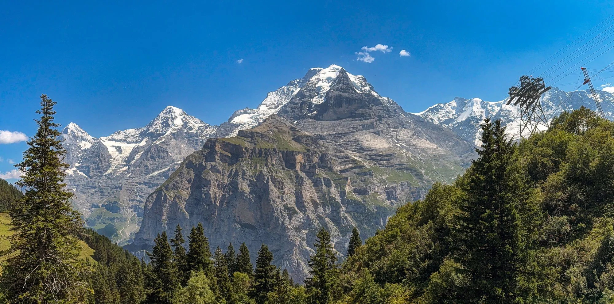 Snow-capped mountain peaks behind green forested hills, clear blue sky, and power lines on the right side.