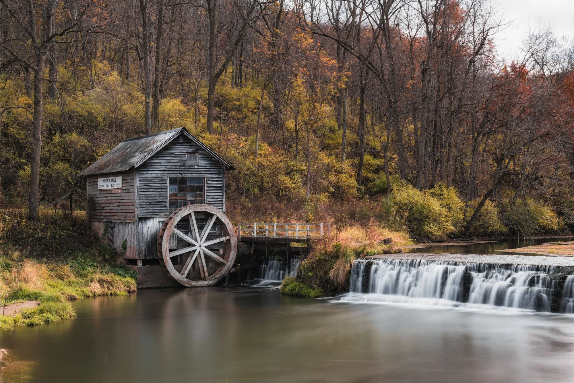 An old wooden watermill beside a flowing creek with small waterfalls, surrounded by autumn trees with orange and yellow leaves.