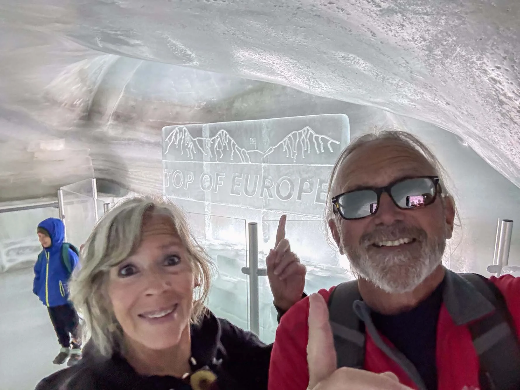 A smiling couple taking a selfie in an ice cave, with a sign that reads "Top of Europe" and contains mountain engravings in the background. A young boy in a blue jacket is visible in the background.
