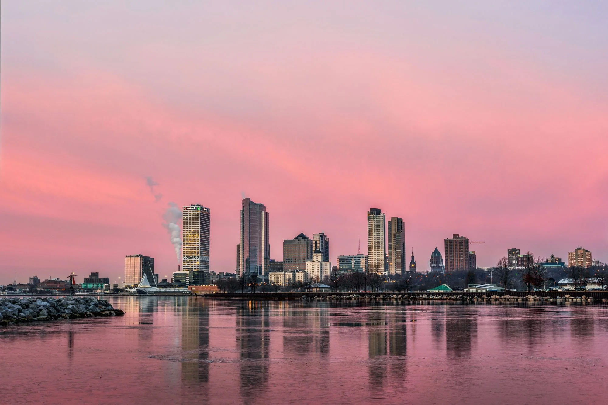 City skyline with tall buildings and skyscrapers at sunset, reflecting on calm water in the foreground, with pink and purple sky.