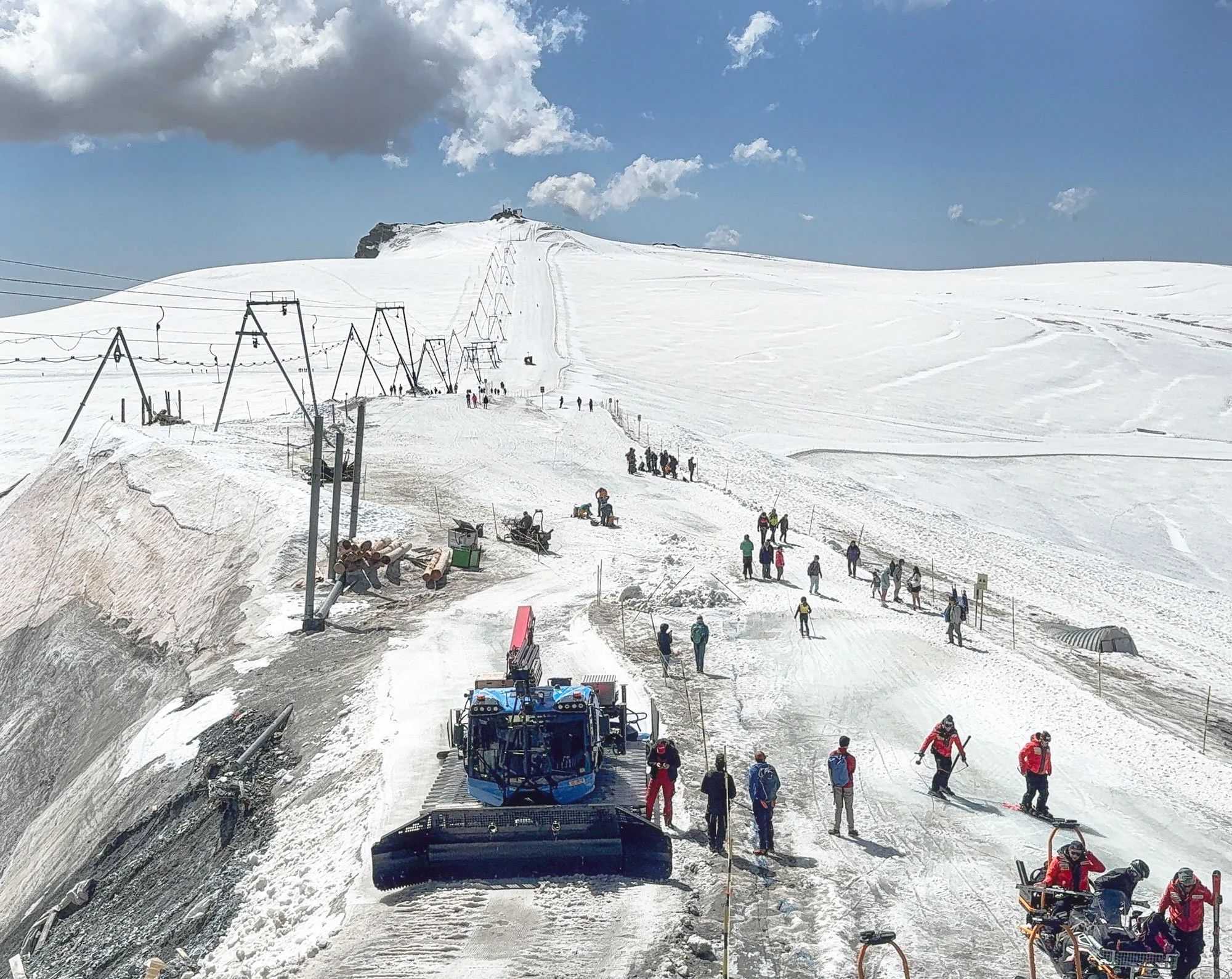 Snow-covered mountain with ski lift poles and cables, people skiing, snowboarding, and walking on the slope, and a snow grooming machine on a paved area at the bottom.