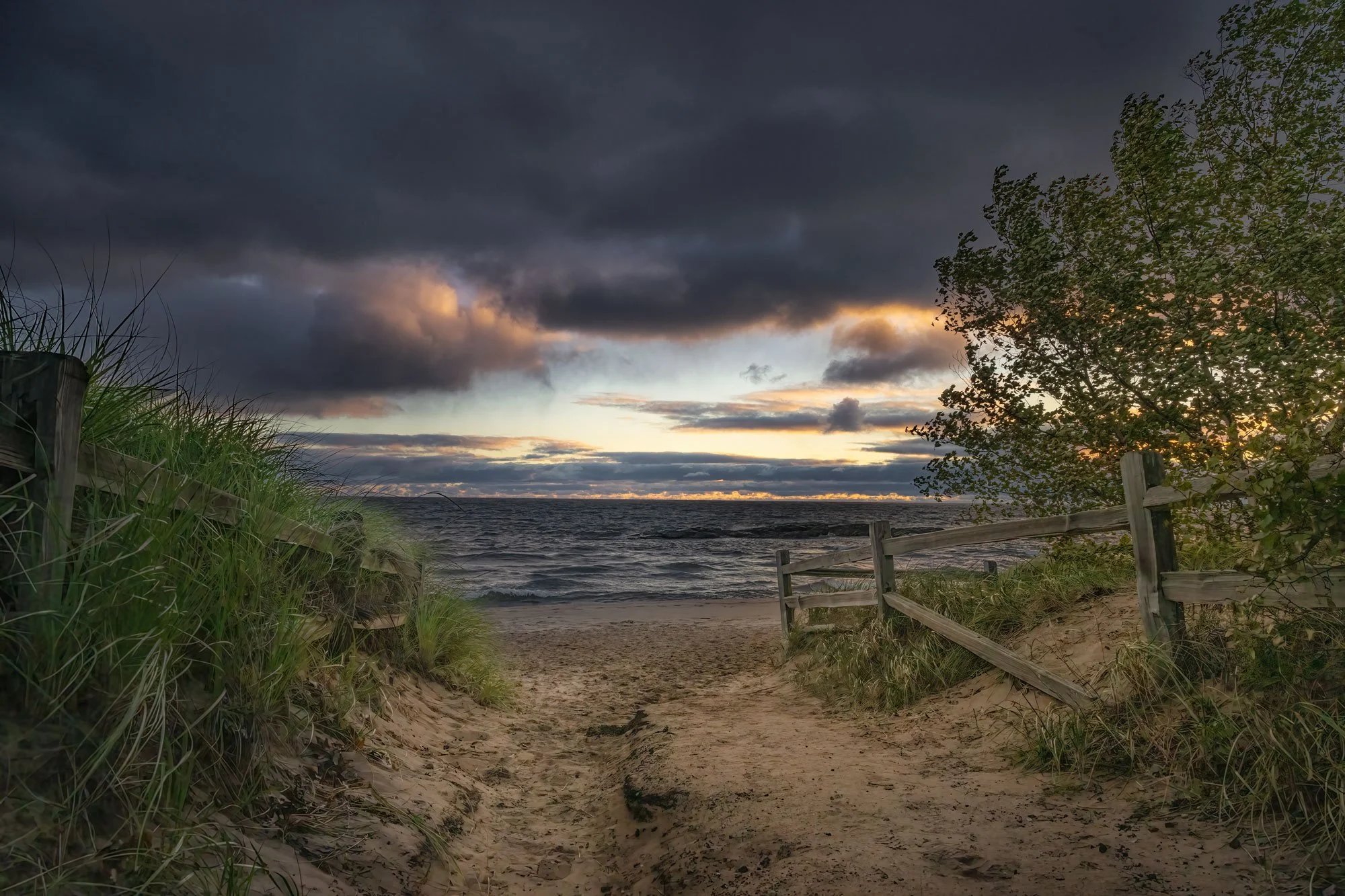 Beach entrance with sandy path, wooden fence, grassy dunes, and beach foliage leading to the ocean under a cloudy sky at sunset.