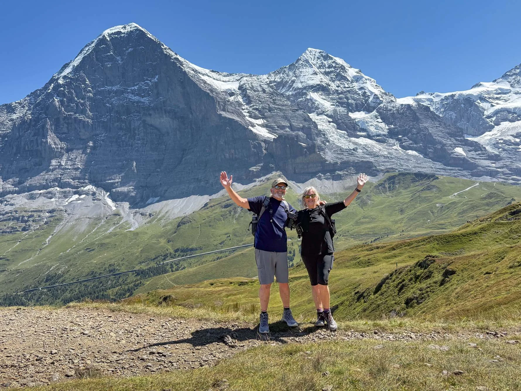 A smiling elderly couple wearing hiking gear standing on a trail in front of a large snow-capped mountain, with green slopes and clear blue sky.