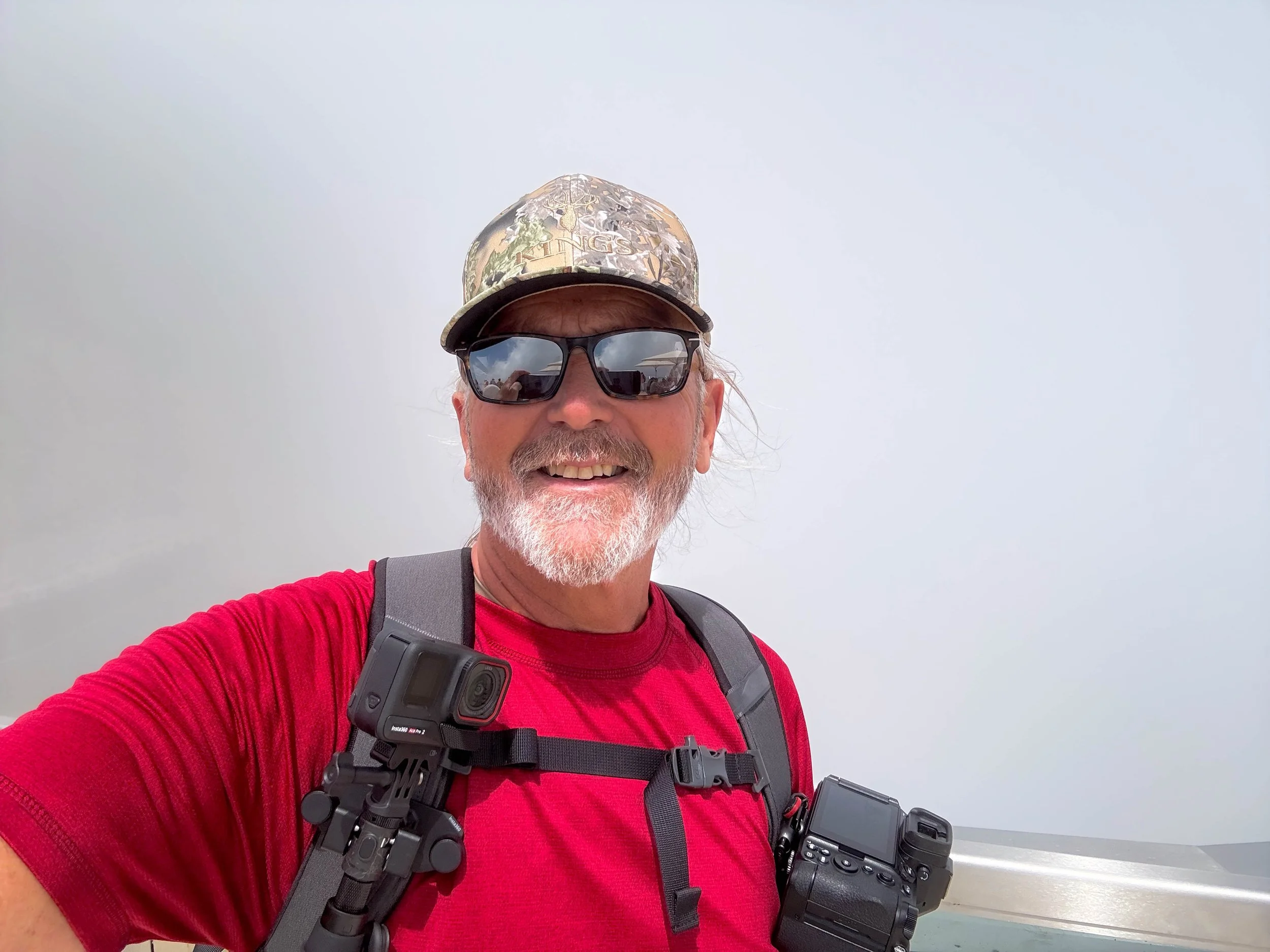 A man with a beard, wearing sunglasses, a camouflage cap, a red shirt, and a backpack with a camera, taking a selfie in a foggy outdoor setting.