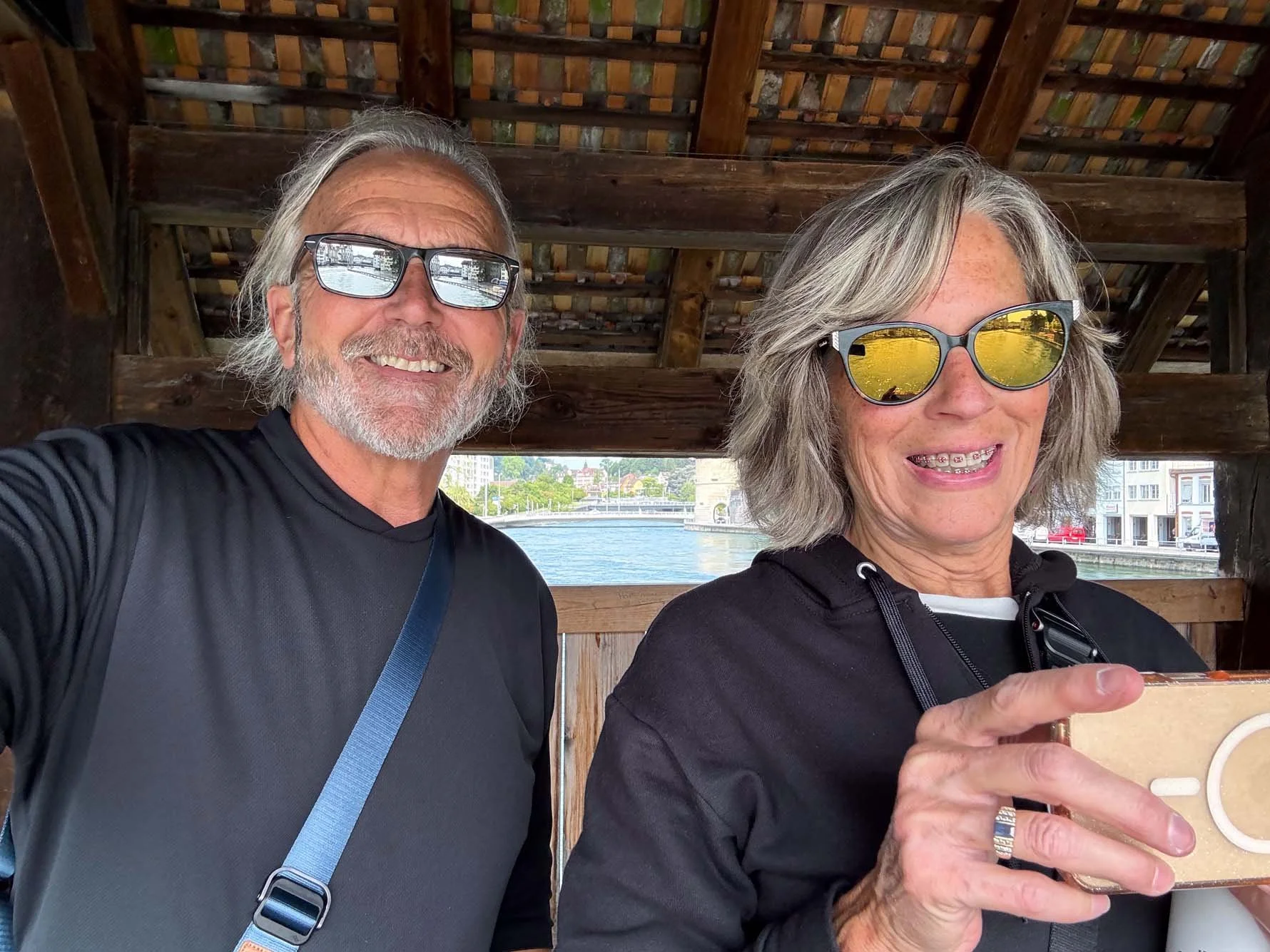 A smiling older man with gray hair, beard, and sunglasses taking a selfie with a smiling older woman with gray hair and sunglasses. They are under a wooden structure near a river or canal, with buildings and boats visible in the background.