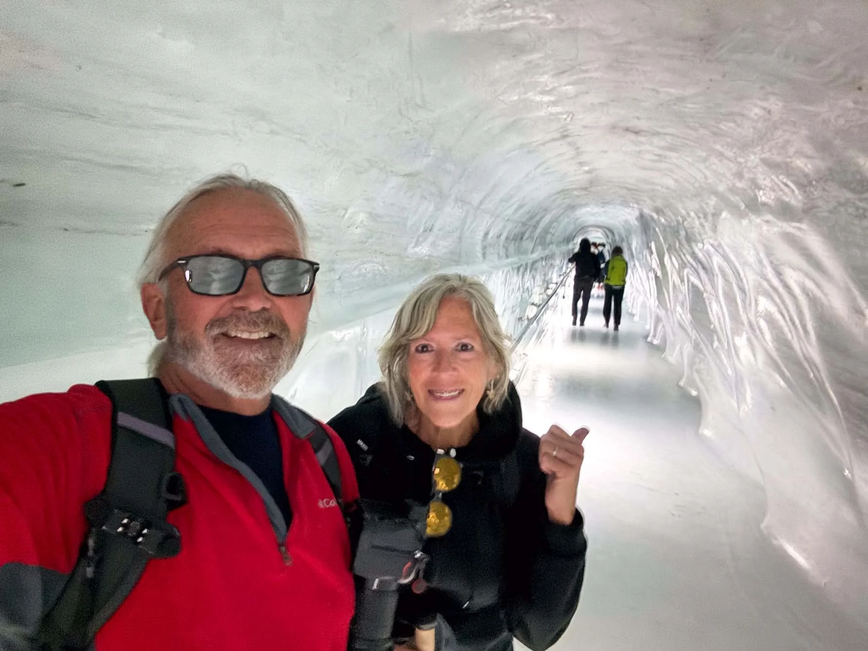 A smiling man with white hair, beard, sunglasses, wearing a red jacket and black backpack, taking a selfie with a woman with short gray hair, smiling, wearing a black jacket, in a snow tunnel with shiny, icy walls. In the background, two people are w
