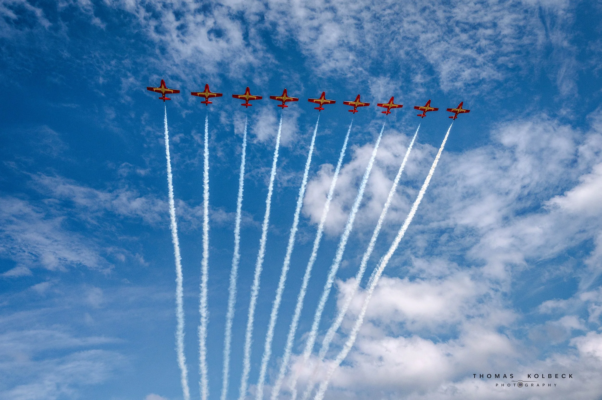 A formation of nine jets flying in a V-shape pattern in the sky, leaving white smoke trails behind them against a backdrop of blue sky with scattered clouds.