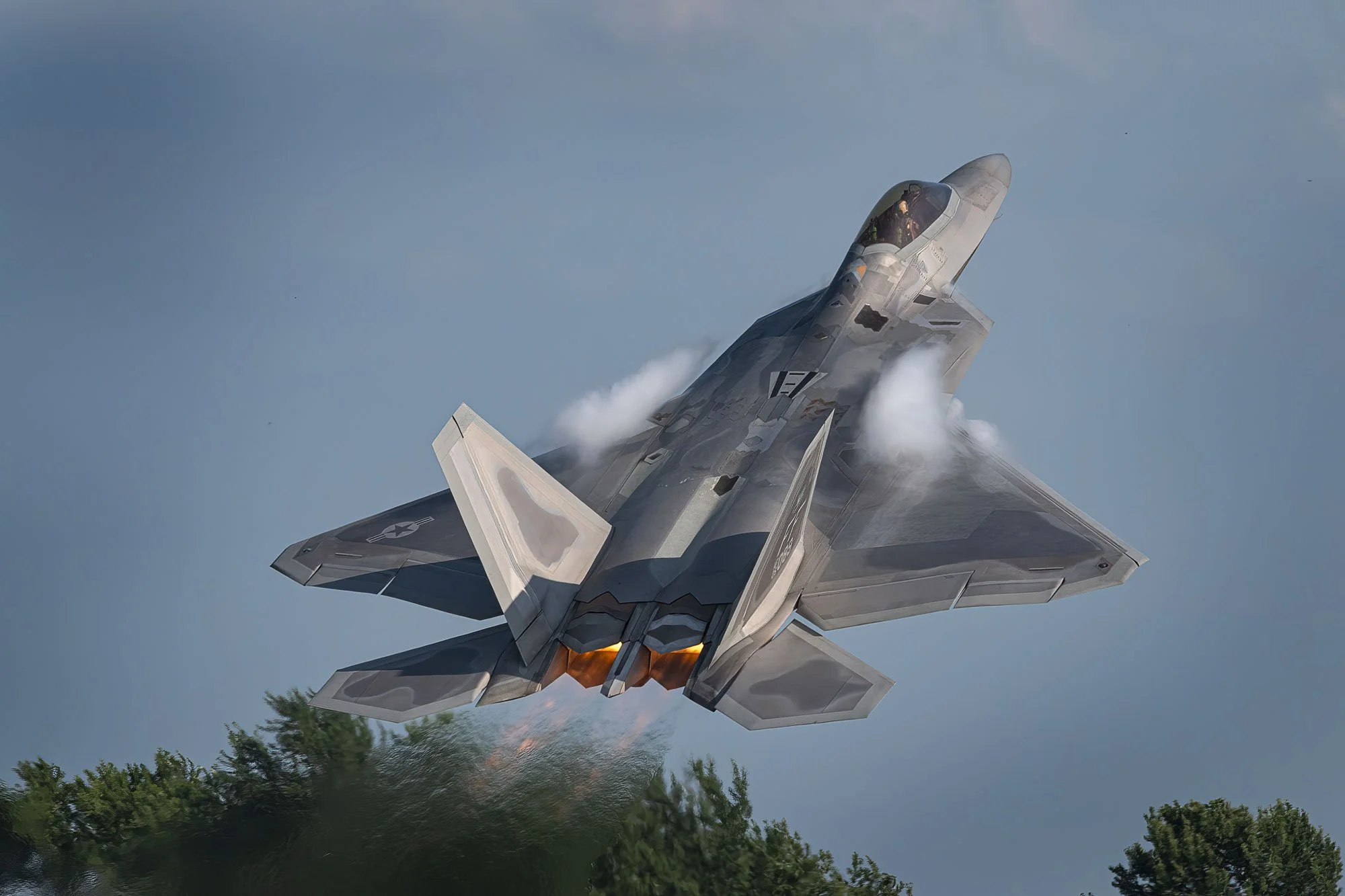 A fighter jet flying at low altitude over trees with afterburners ignited and leaving vapor trails.