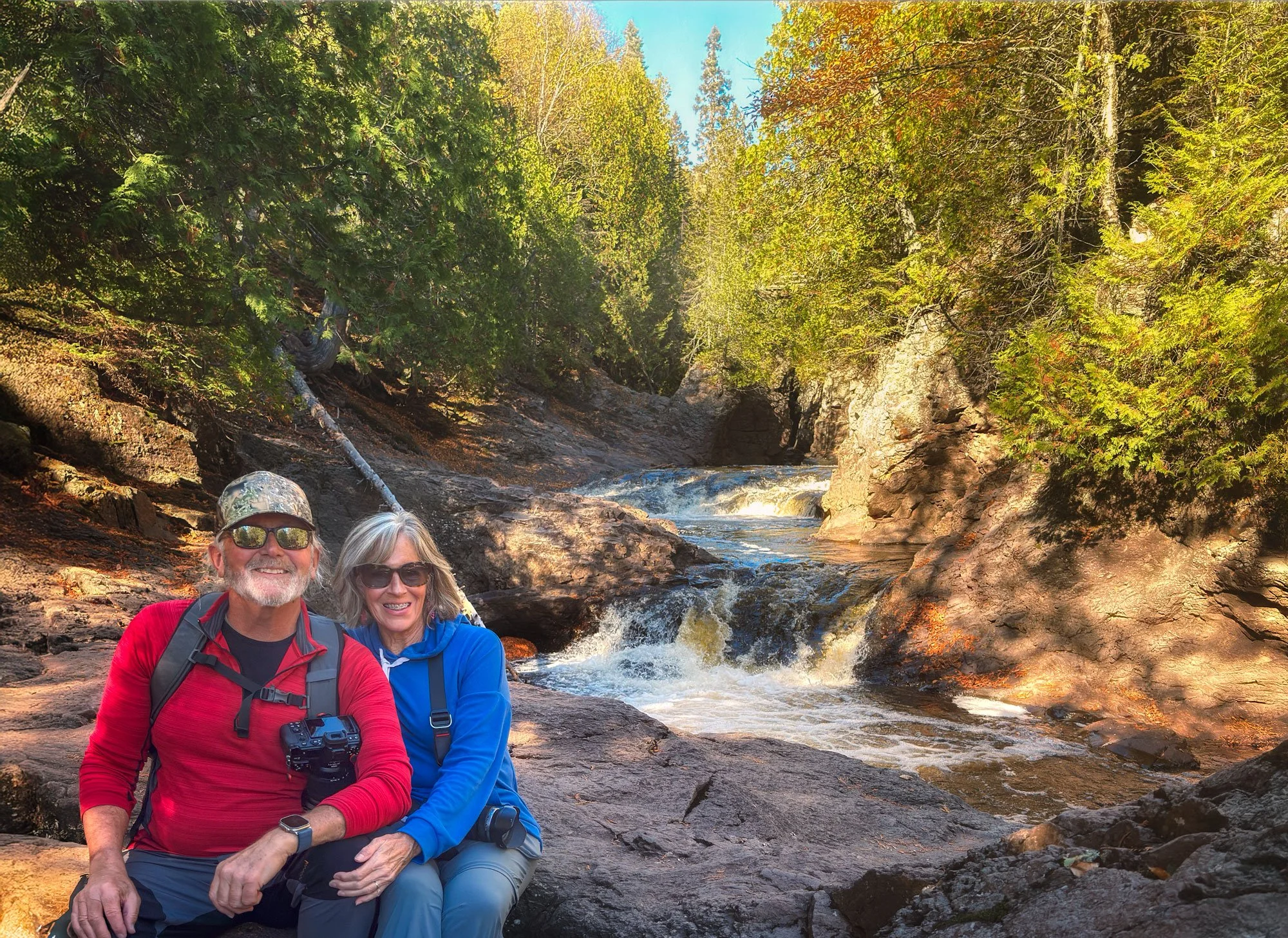 Two smiling hikers, a man in a red jacket and a woman in a blue jacket, sitting on rocks beside a small waterfall and river in a forested area during daytime.
