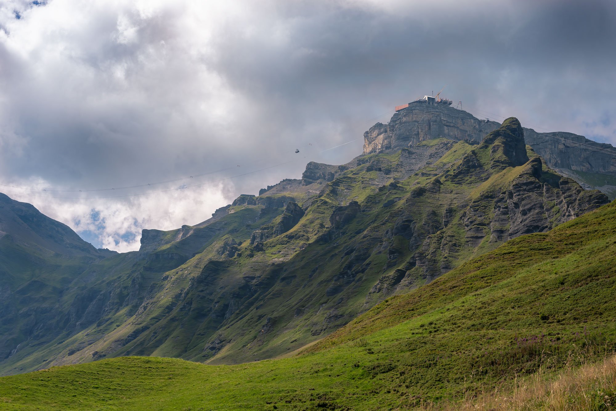 A mountain with a cable car and station on top, green grassy slopes, cloudy sky.