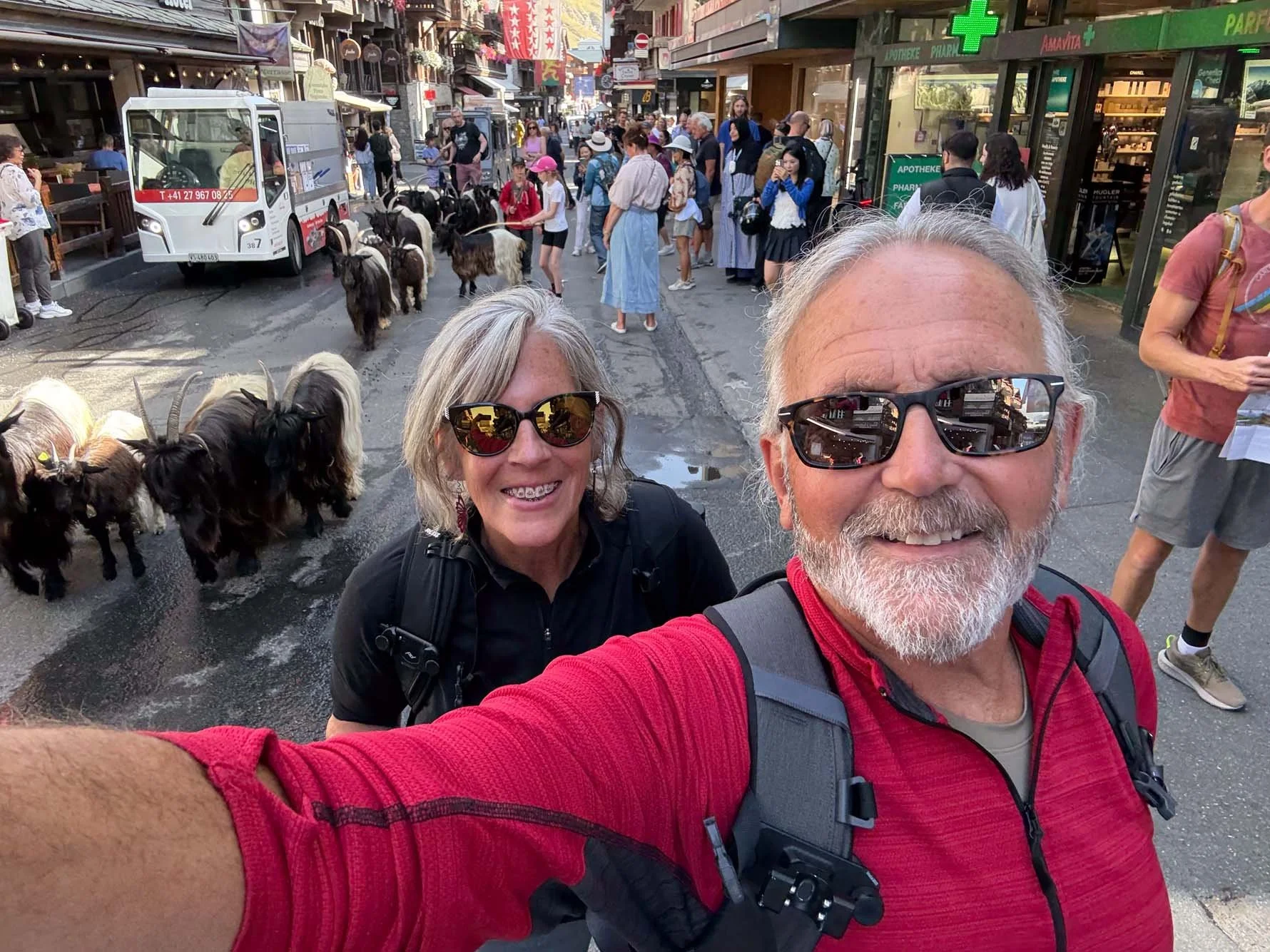 Smiling man and woman taking selfie on busy street filled with tourists, goats, and storefronts, including a pharmacy and a garbage truck.