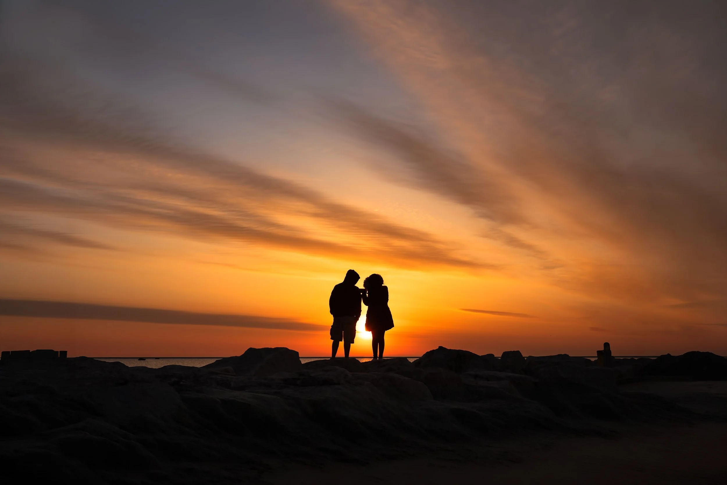 Silhouettes of a couple standing on rocks near the beach at sunset, with colorful sky and clouds in the background.