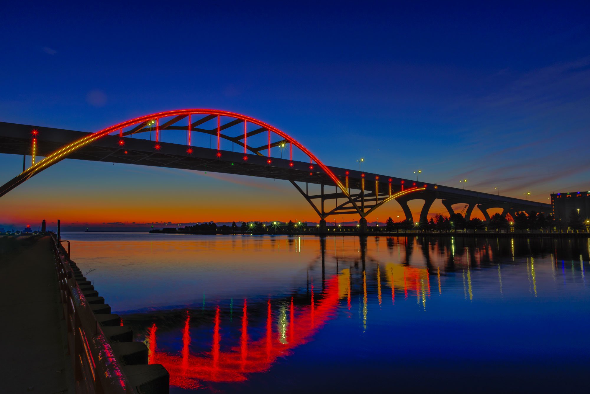 A bridge illuminated with red, yellow, and green lights stands over a body of water during sunset, with the sky displaying shades of blue and orange and the bridge's reflection visible in the water.