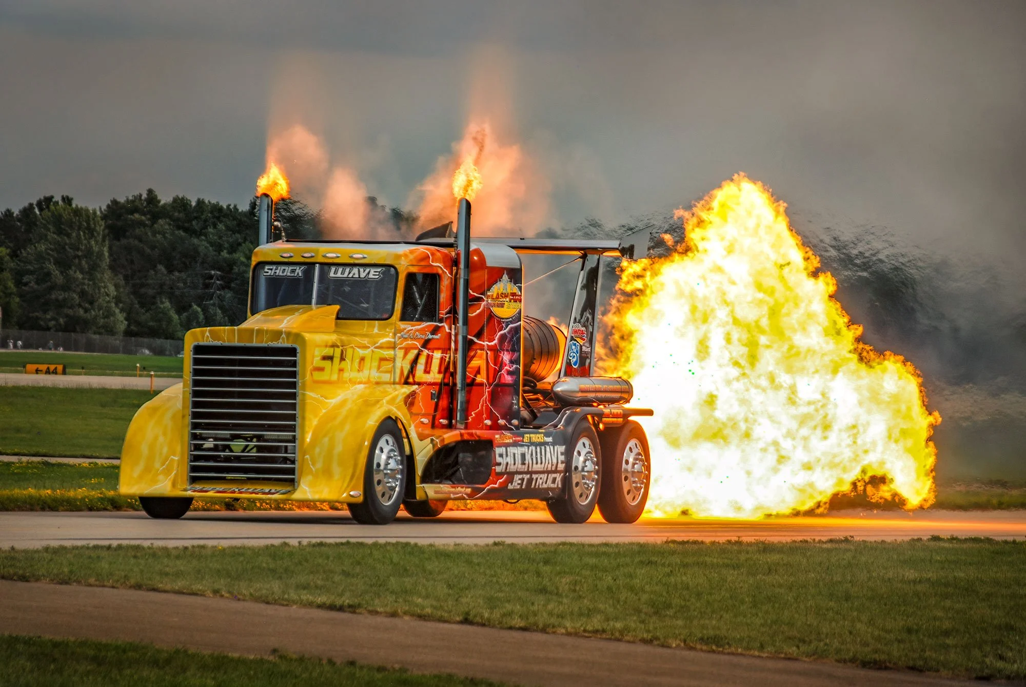 A yellow jet truck named Shockwave emitting flames and smoke during a demonstration or stunt performance.