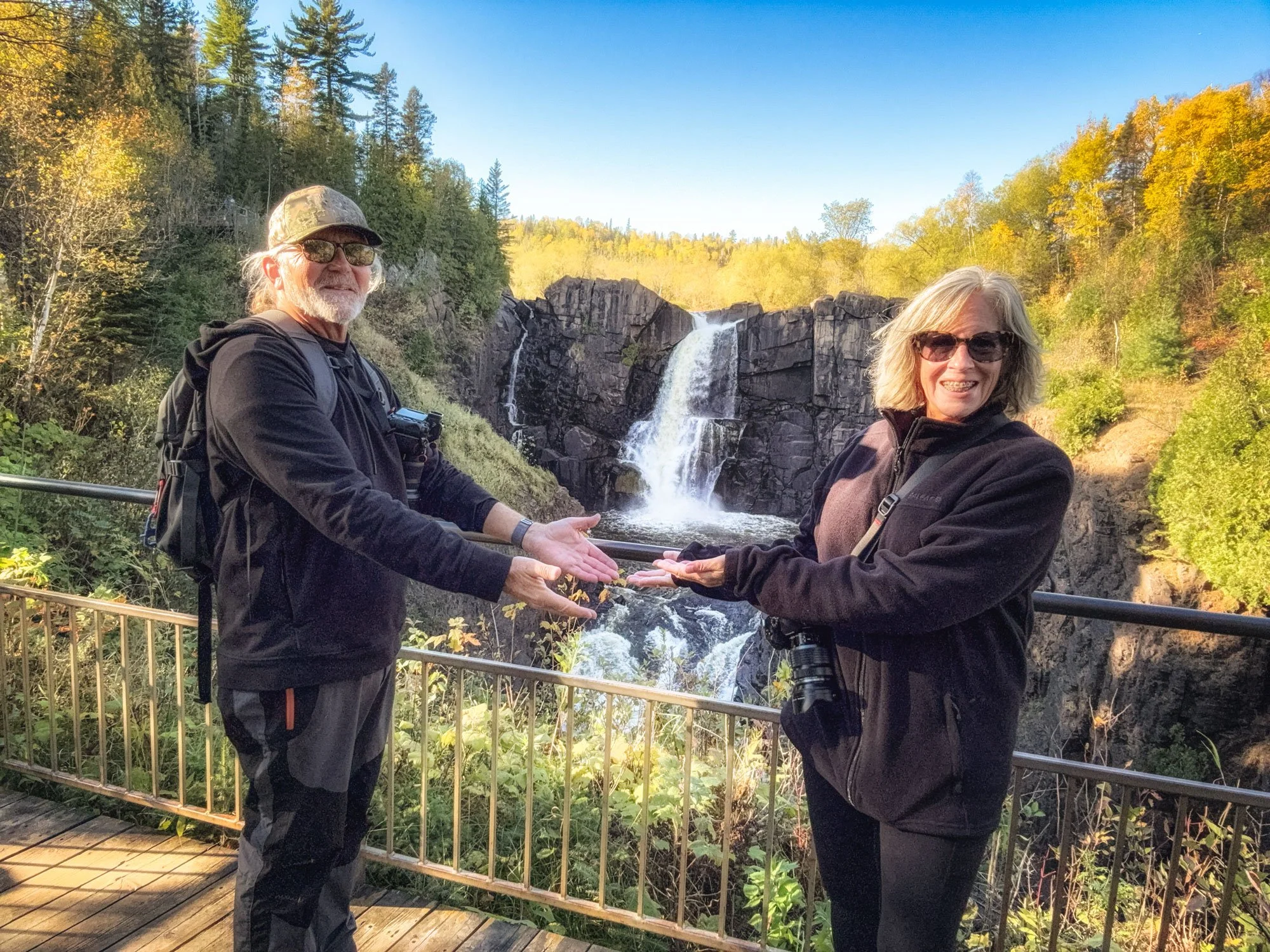 A man and woman standing on a wooden viewing platform with a waterfall and rocky cliff in the background. The man has long hair, a beard, sunglasses, and a backpack, and the woman has shoulder-length hair, sunglasses, and a camera hanging from her ne