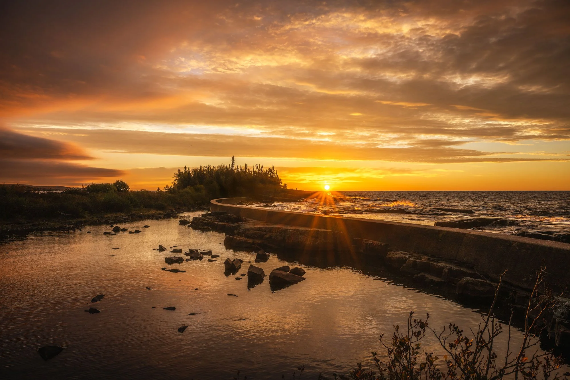 Sunset over the ocean with a curved walkway, rocks in the water, and cloudy sky.