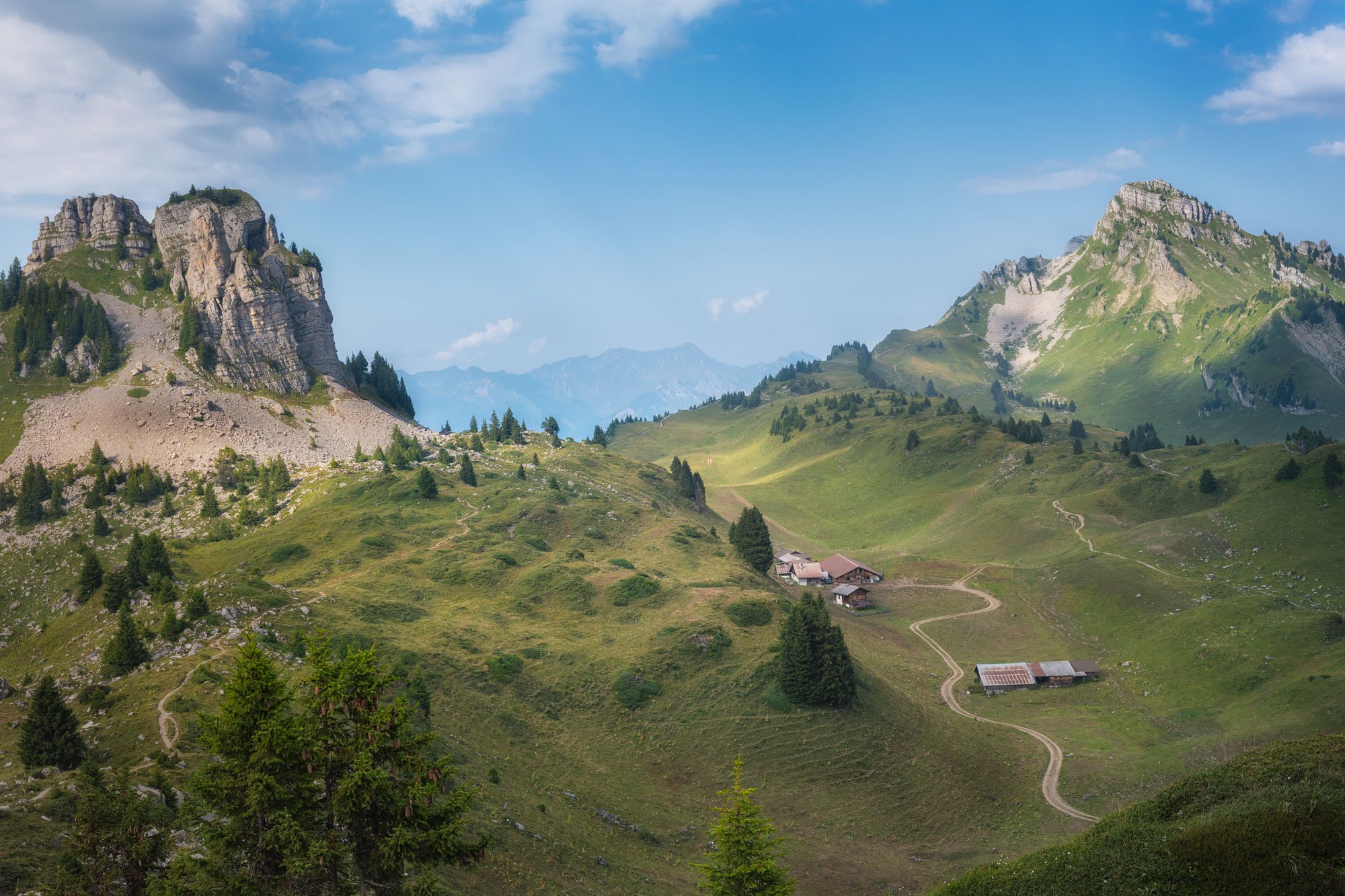 Lush green mountains with rocky peaks and a winding dirt road leading to small houses, under a partly cloudy sky.