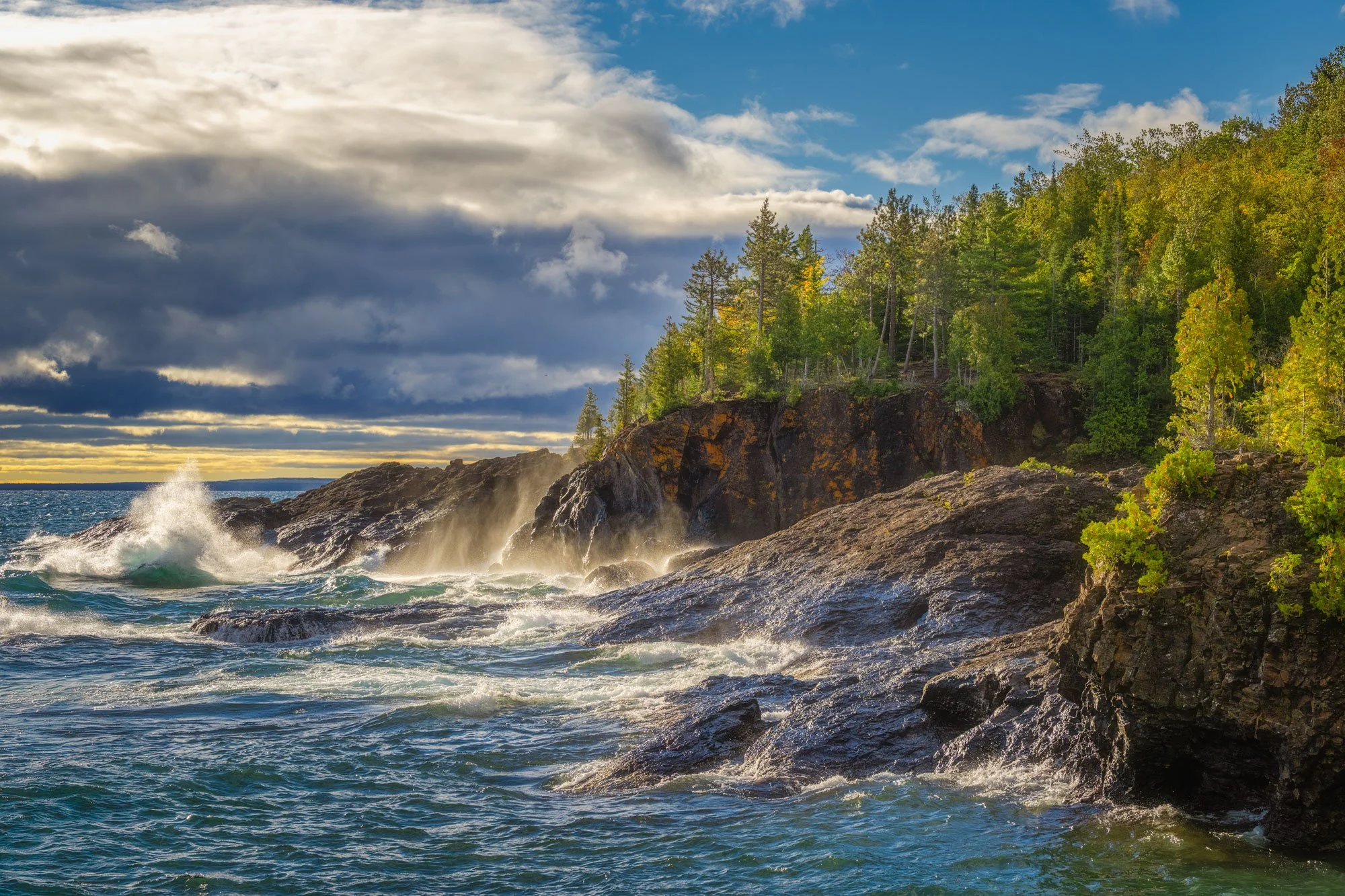 Waves crashing against rocky shoreline with green trees on a cliff, under partly cloudy sky with sunlight peeking through.