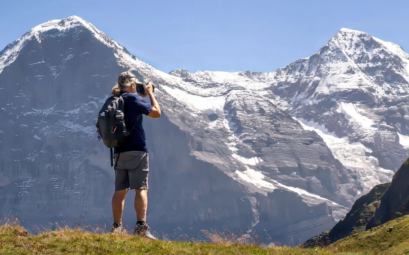 A hiker taking a photo of snow-capped mountains , carrying a backpack, on a grassy patch under a clear blue sky in Switzerland.