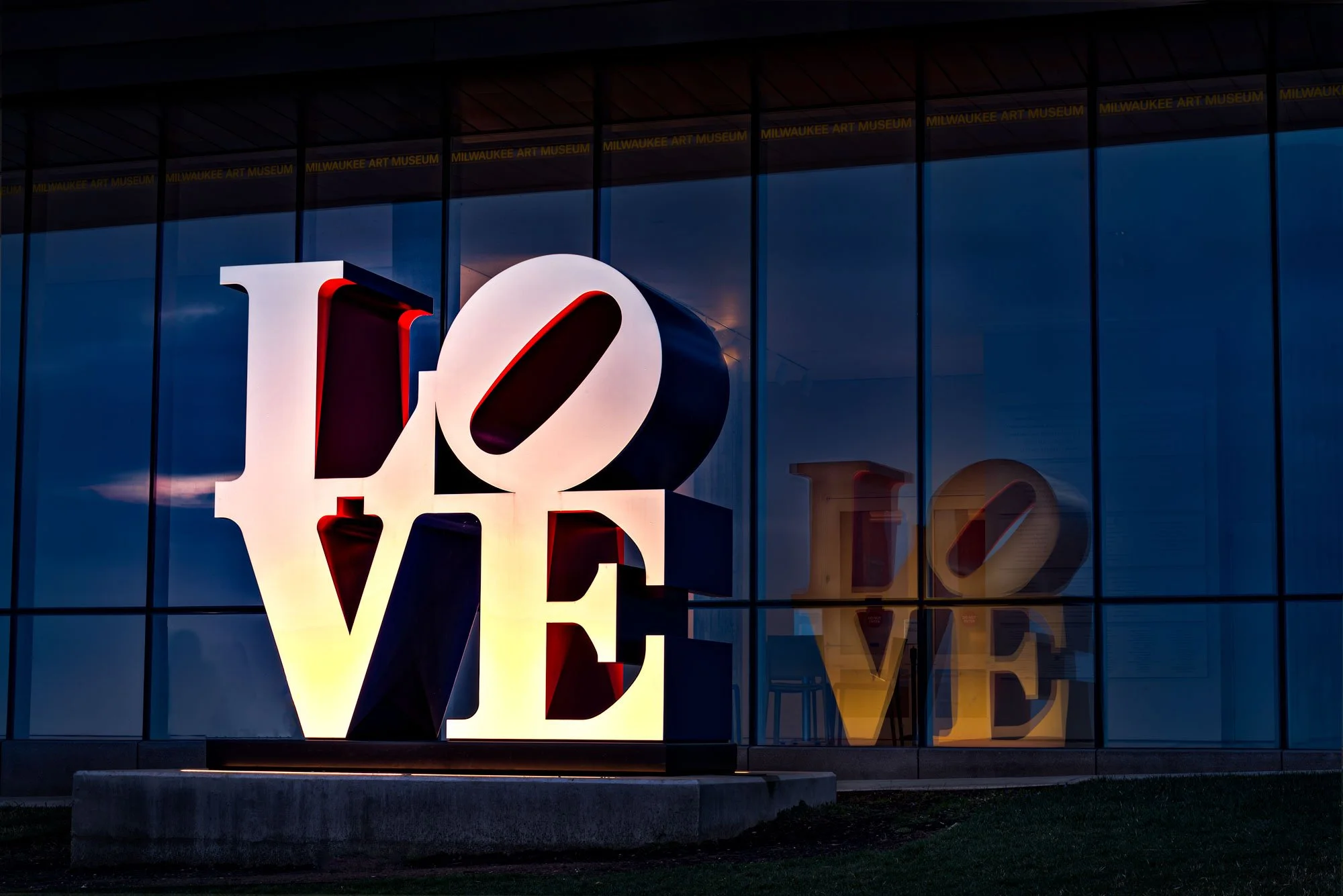 Large, illuminated 'LOVE' sculpture outside the Milwaukee Art Museum with a glass building in the background reflecting the sign.
