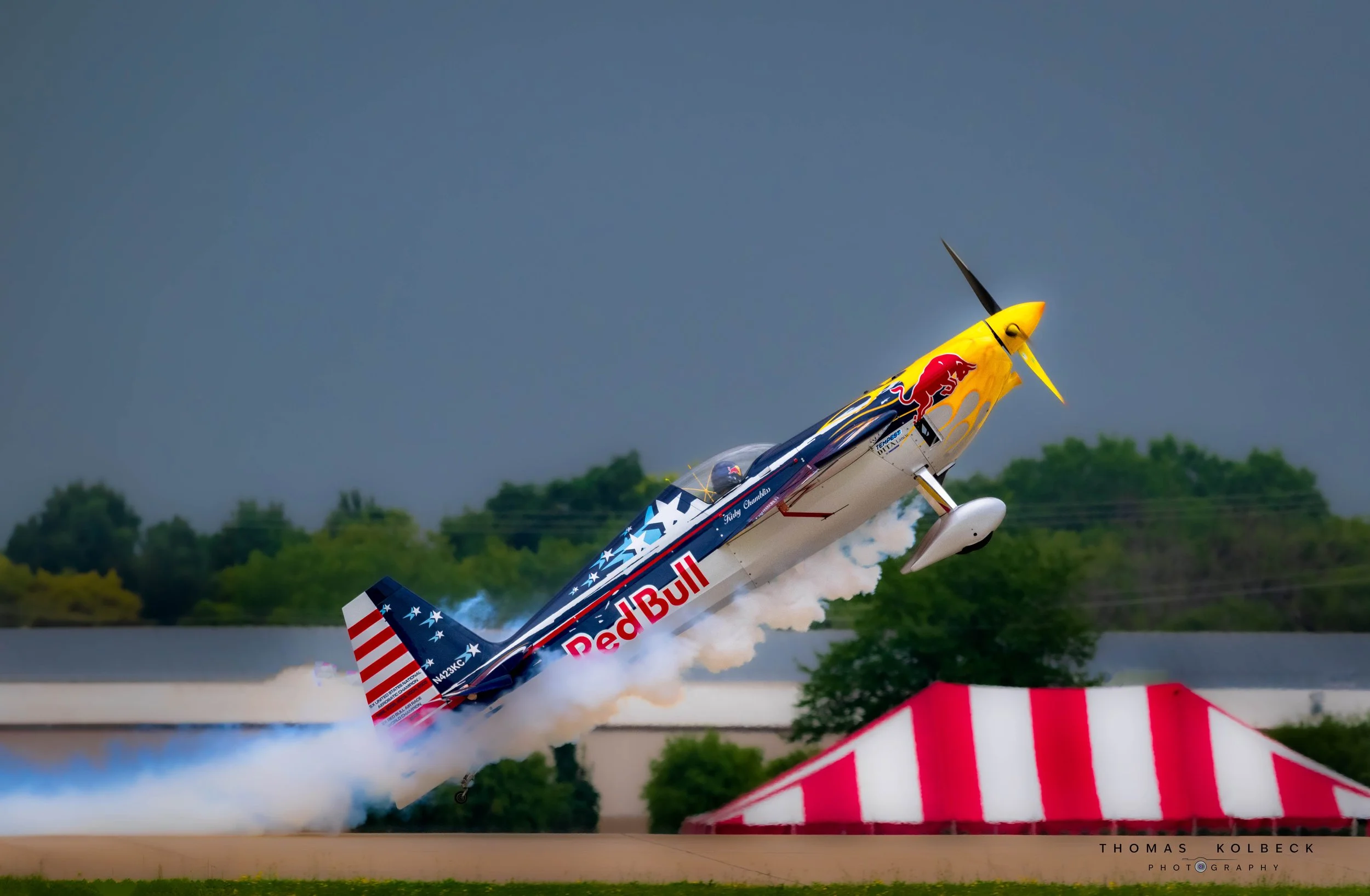 A Red Bull aerobatic aircraft taking off, leaving a trail of smoke with a blue sky and trees in the background.