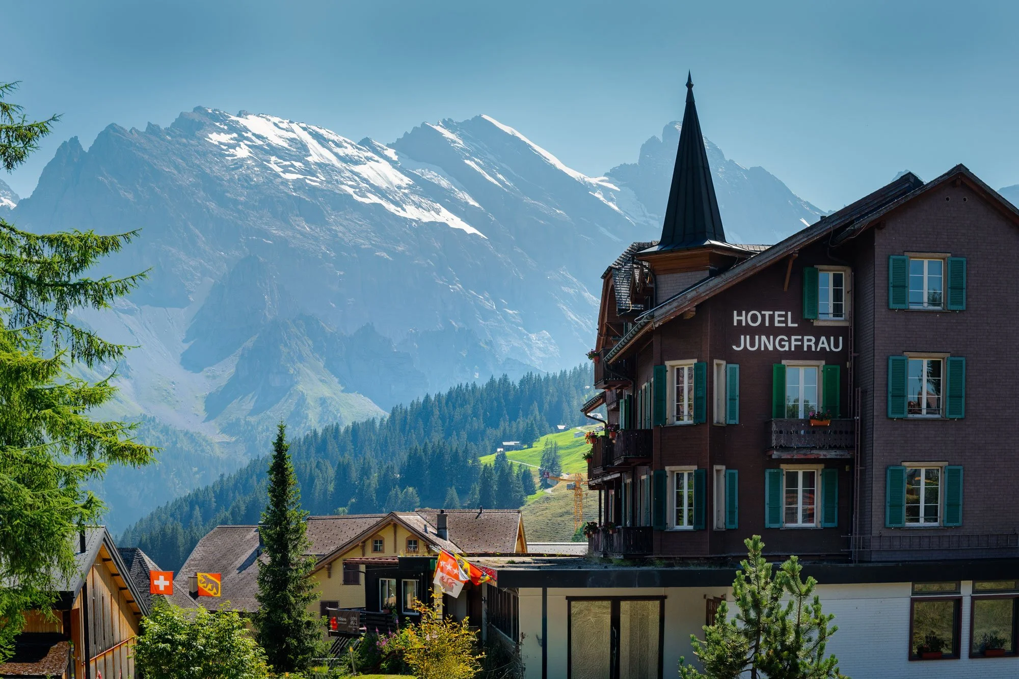 A mountain town with a hotel named 'Hotel Jungfrau' in the foreground, surrounded by lush trees and snow-capped peaks in the background.