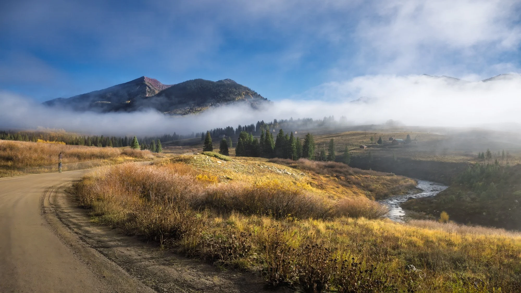 Scenic view of a mountainous landscape with a dirt road in the foreground, rolling hills covered with grass and trees, a small stream, and mountains partly obscured by clouds and fog under a blue sky.