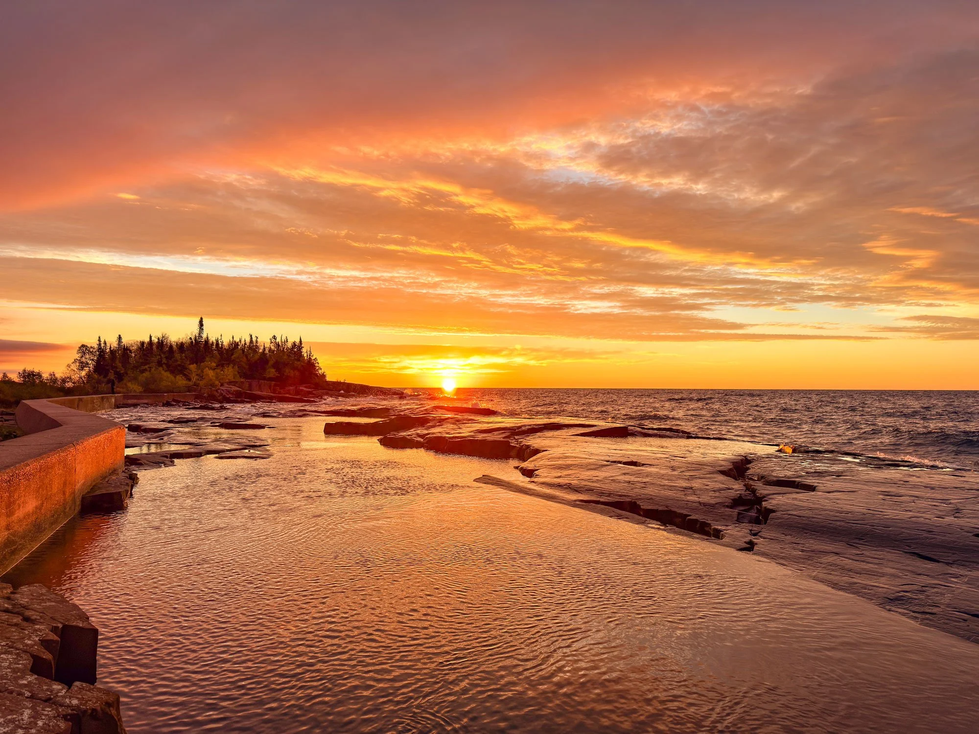 Sunset over rocky shoreline with trees in the background, reflecting the colorful sky on the water.