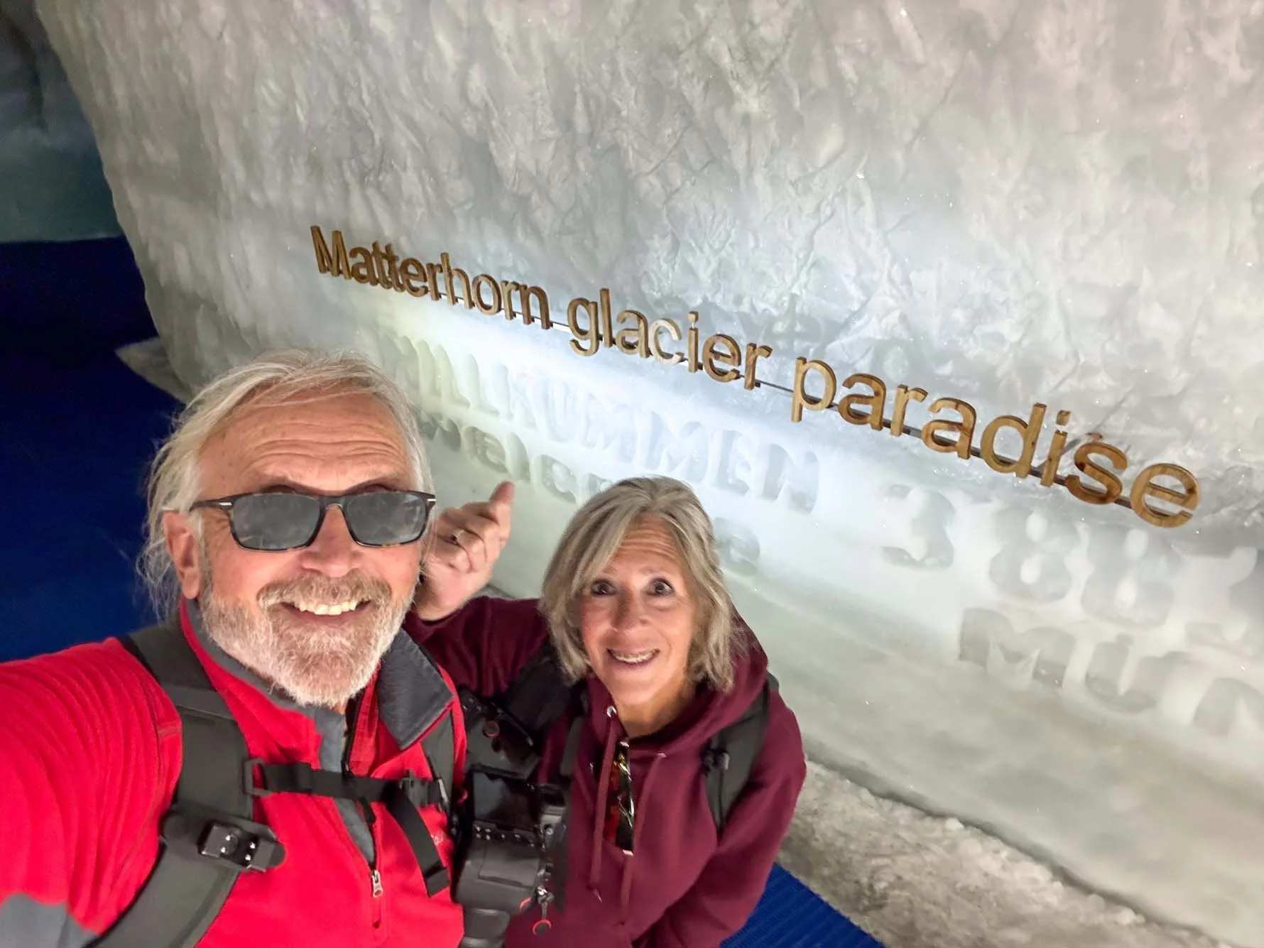 An older man and woman taking a selfie inside Matterhorn Glacier Paradise, with a large ice wall behind them displaying the location name.