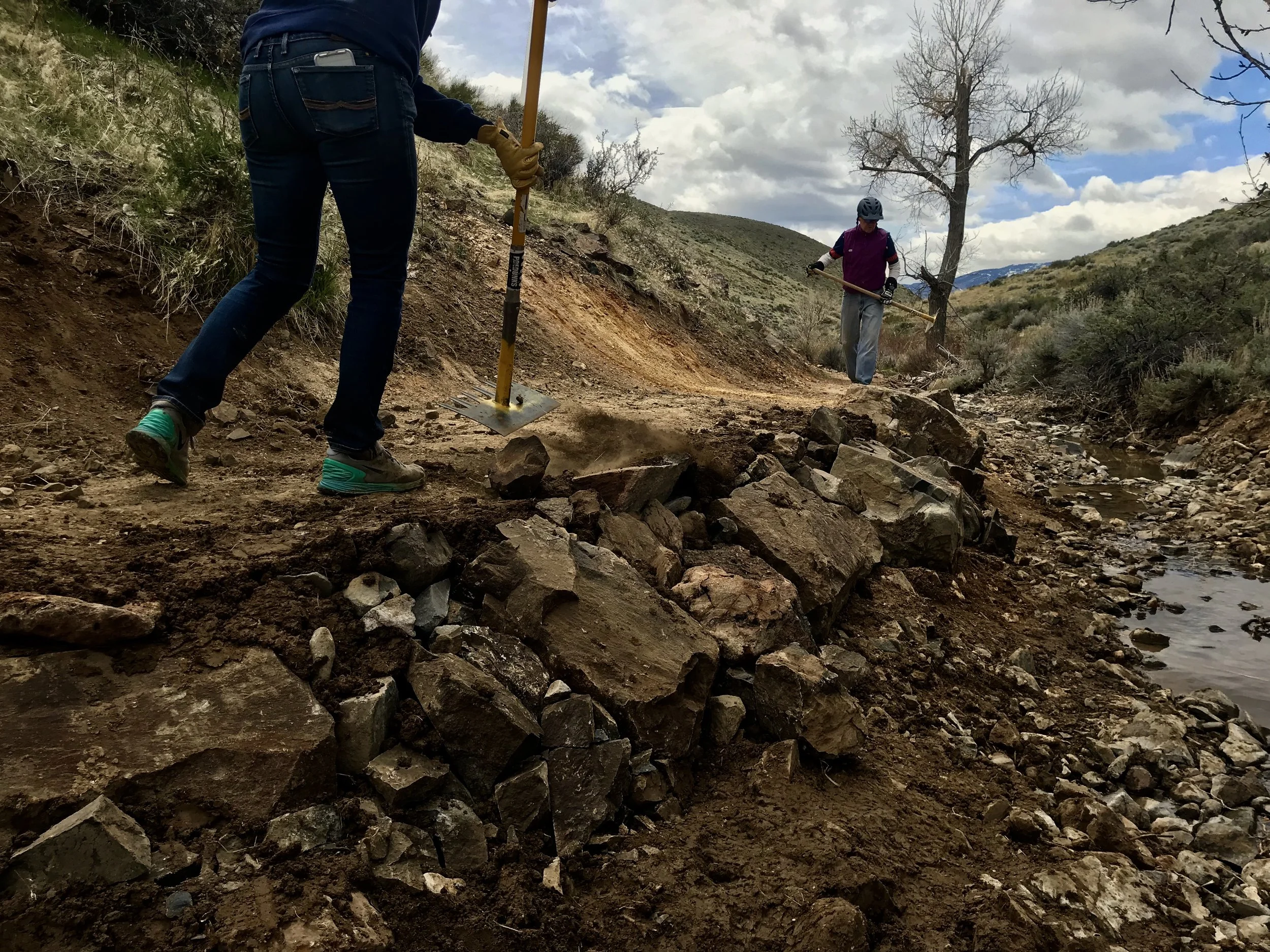 Two people working on a stream restoration project by placing rocks along the stream bank during the daytime. One person in the foreground is using a shovel, and another in the background is holding a tool.