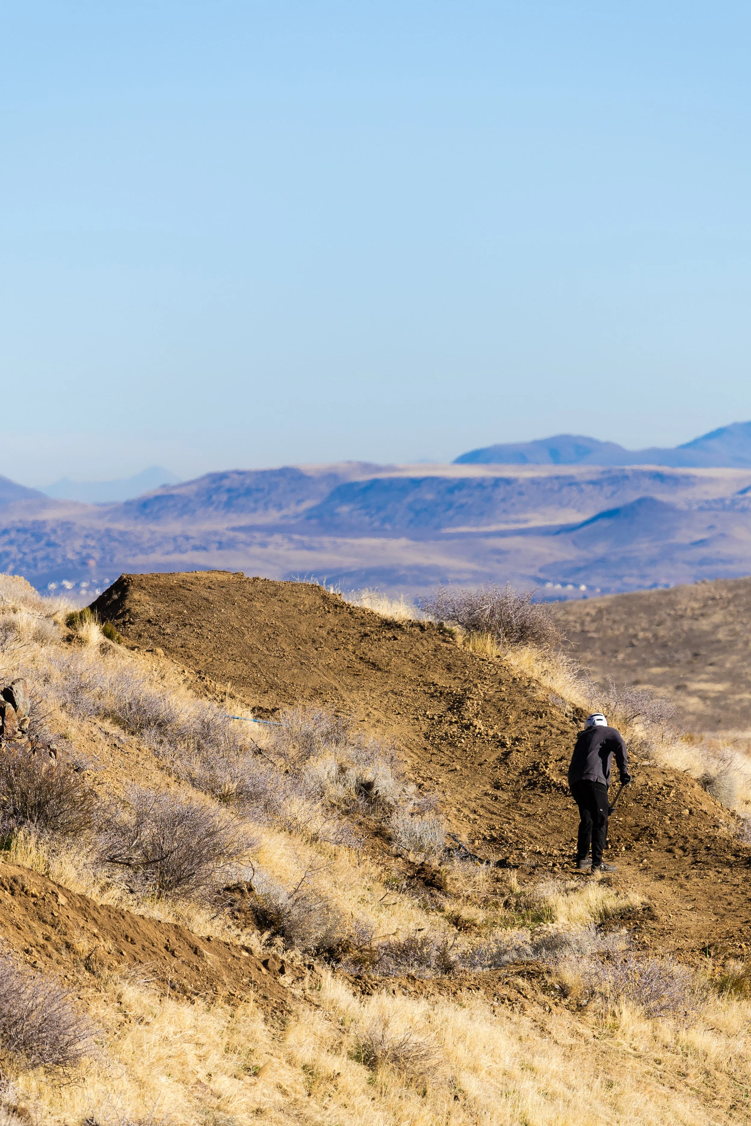 A person wearing black outdoor clothing and a white helmet is climbing uphill on a dirt trail in a desert landscape with dry grass and shrubs, with mountains in the background under a clear blue sky.