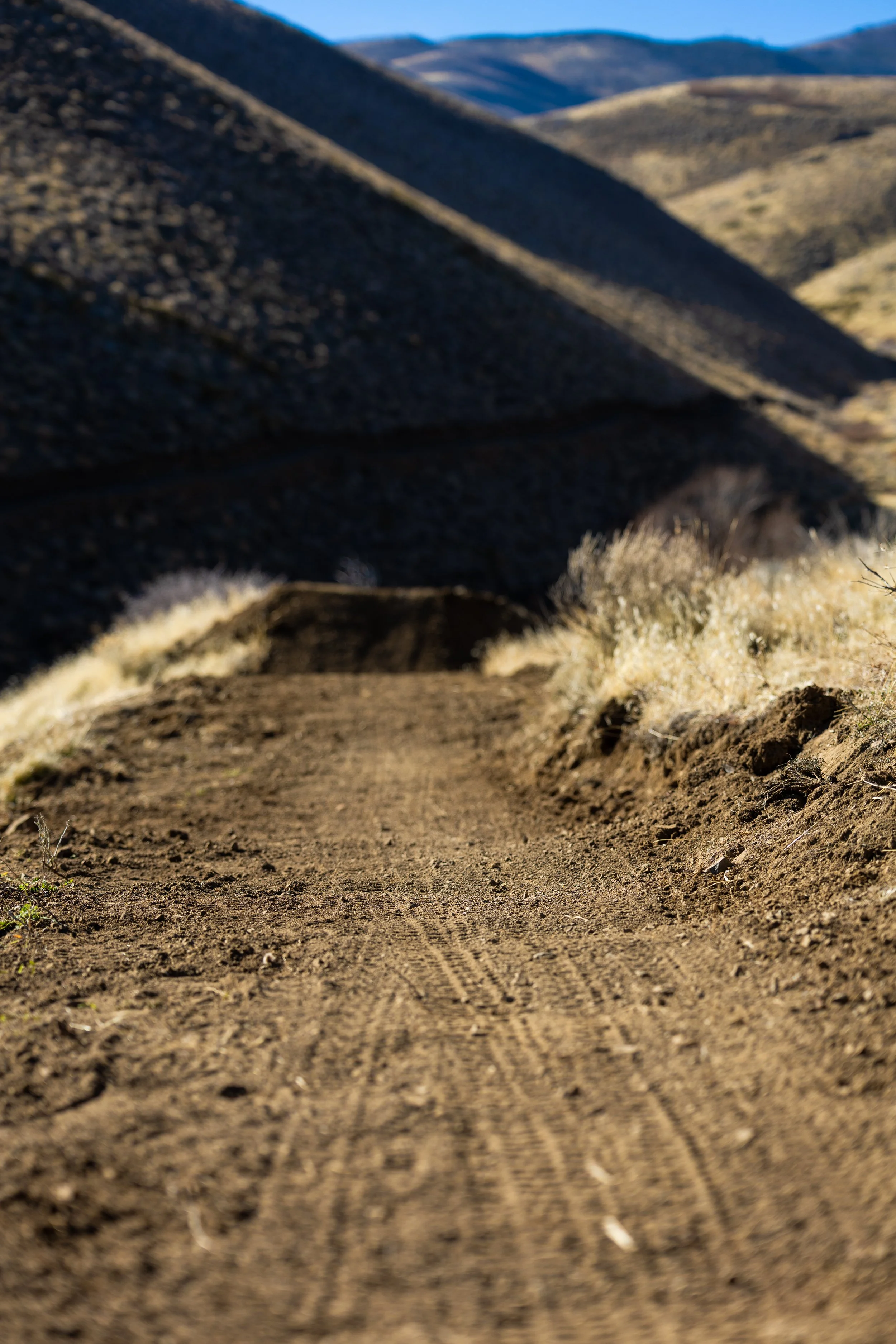 Dirt trail through a mountainous desert landscape with dry grass and rocky hills in the background.