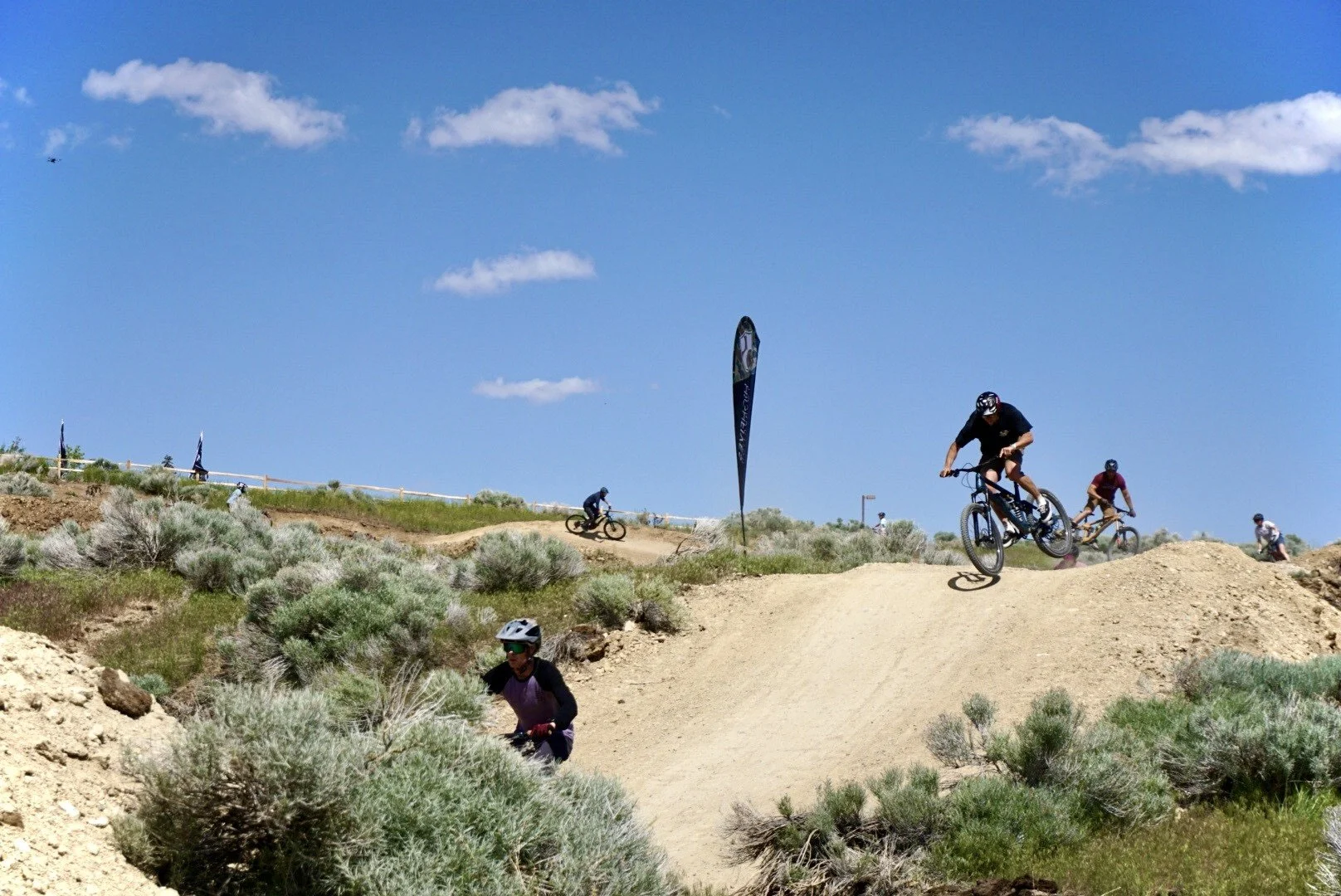 Two mountain bikers riding on a dirt trail in a desert landscape with dry bushes and clear blue sky.