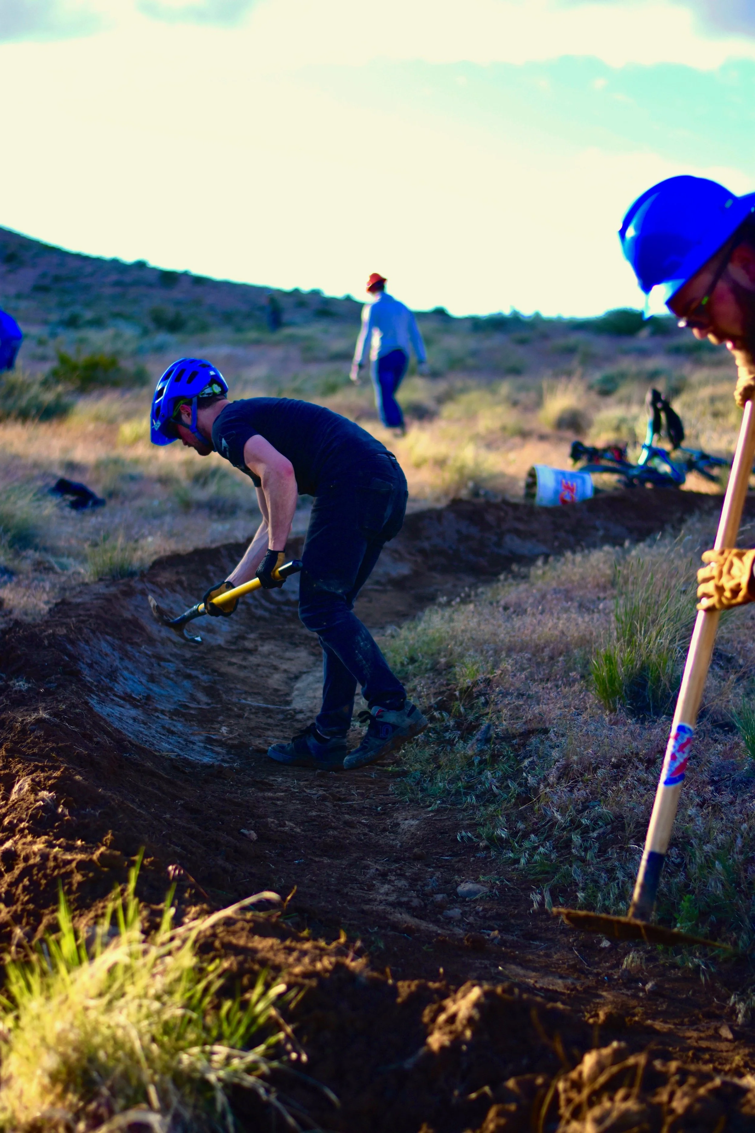 People working on a dirt trail in a hilly outdoor area, some wearing safety helmets and gloves, with a shovel, brush, and other tools, during the daytime.