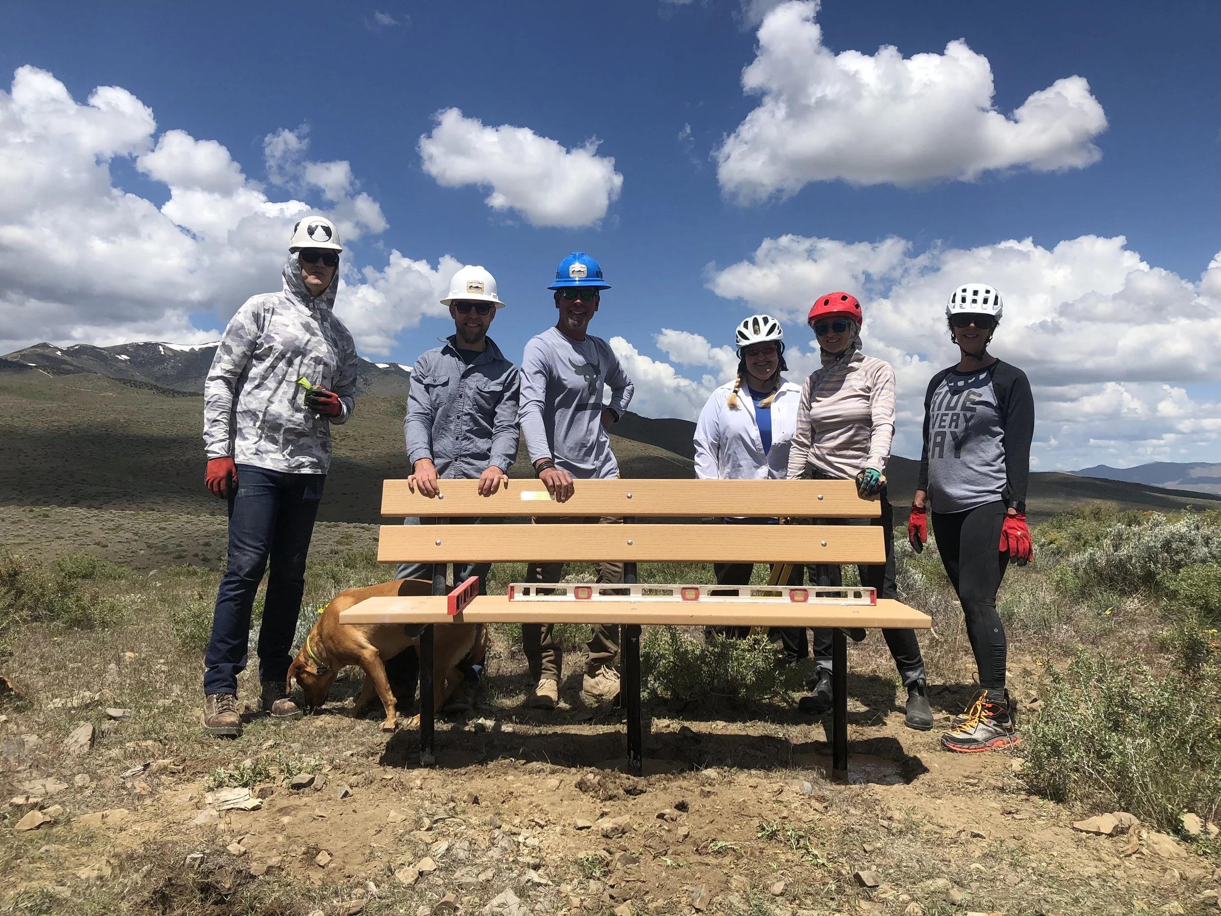 A group of seven people, wearing helmets and gloves, standing on an outdoor trail with mountains in the background, posing with a wooden park bench, along with a dog.