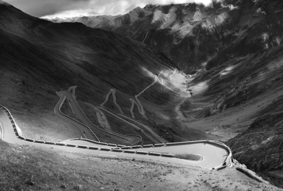 A winding mountain road with sharp turns and guardrails, surrounded by rugged terrain and mountains in the background, in black and white.